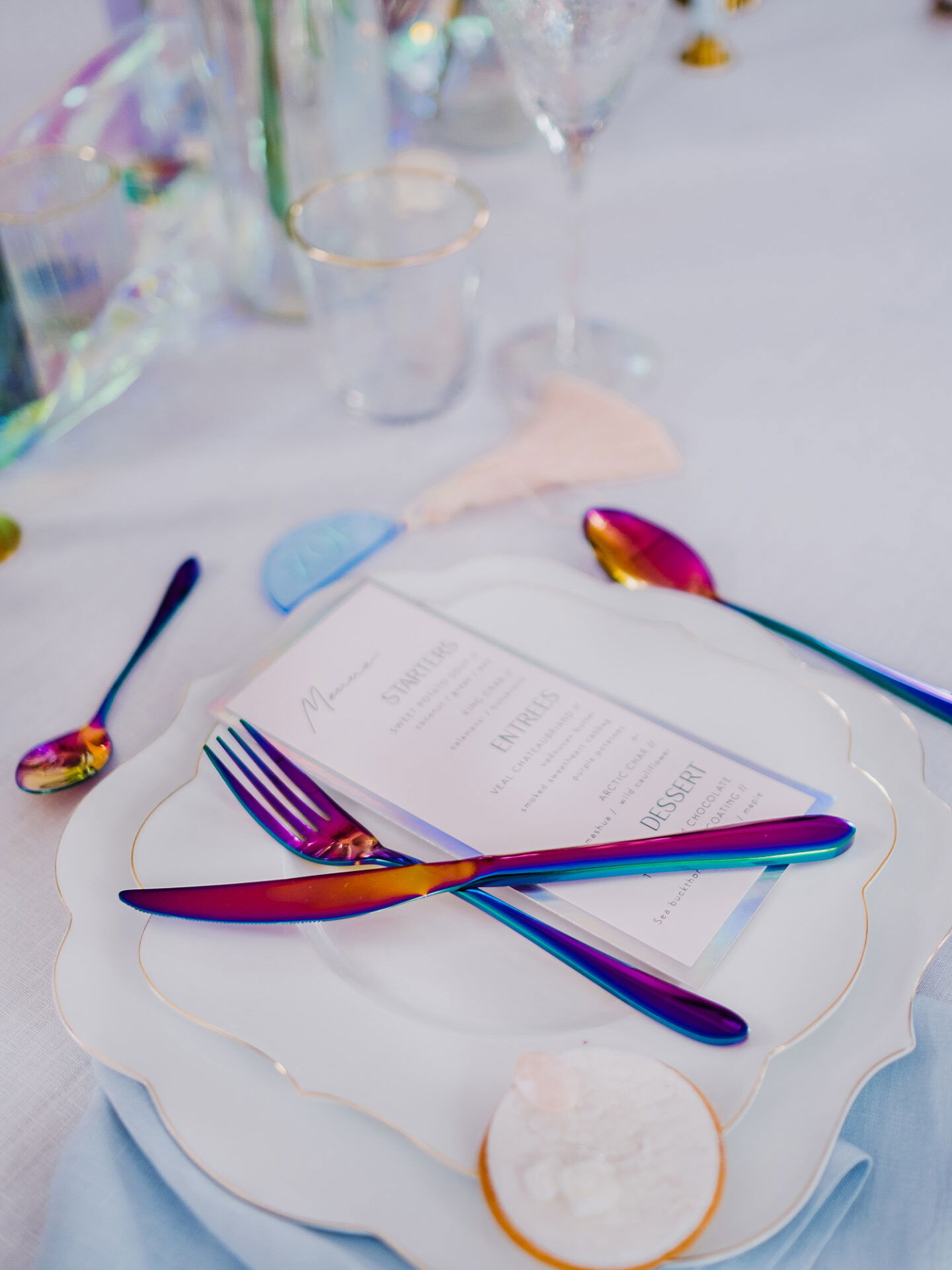 Ein Teller mit Menükarte und schillerndem Besteck in Regenbogenfarben, umgeben von elegantem Glas. Die feinen Goldränder der Teller sowie die pastellfarbenen Accessoires verleihen der Tischdekoration eine stilvolle und moderne Eleganz.