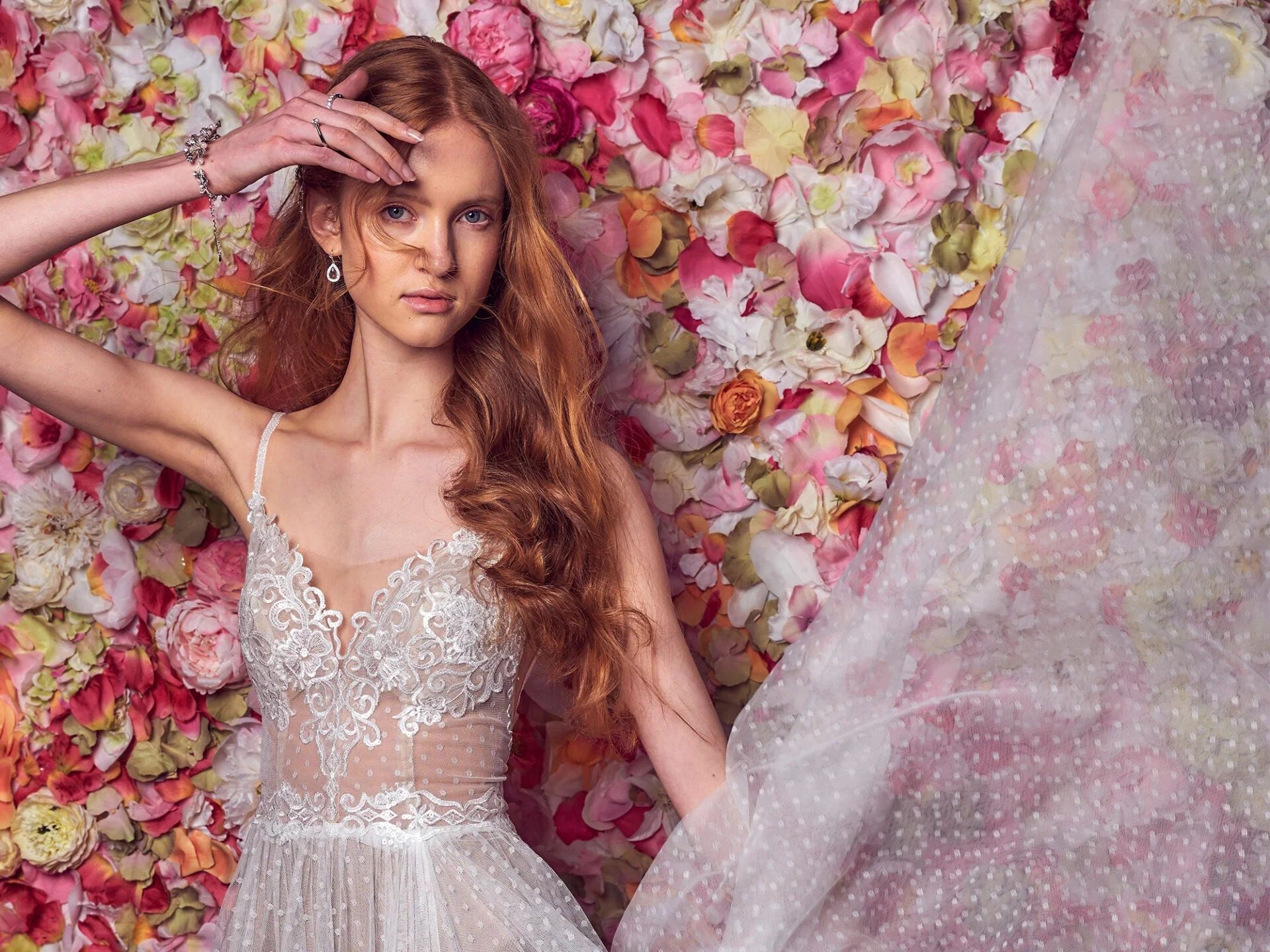 Eine junge Braut posiert in einem voluminösen weißen Kleid auf einem antiken Stuhl. Hinter ihr eine Wand voller Ranunkeln in zarten Rosa- und Cremetönen, die das großflächige Kleid umrahmen und eine märchenhafte, florale Kulisse schaffen.