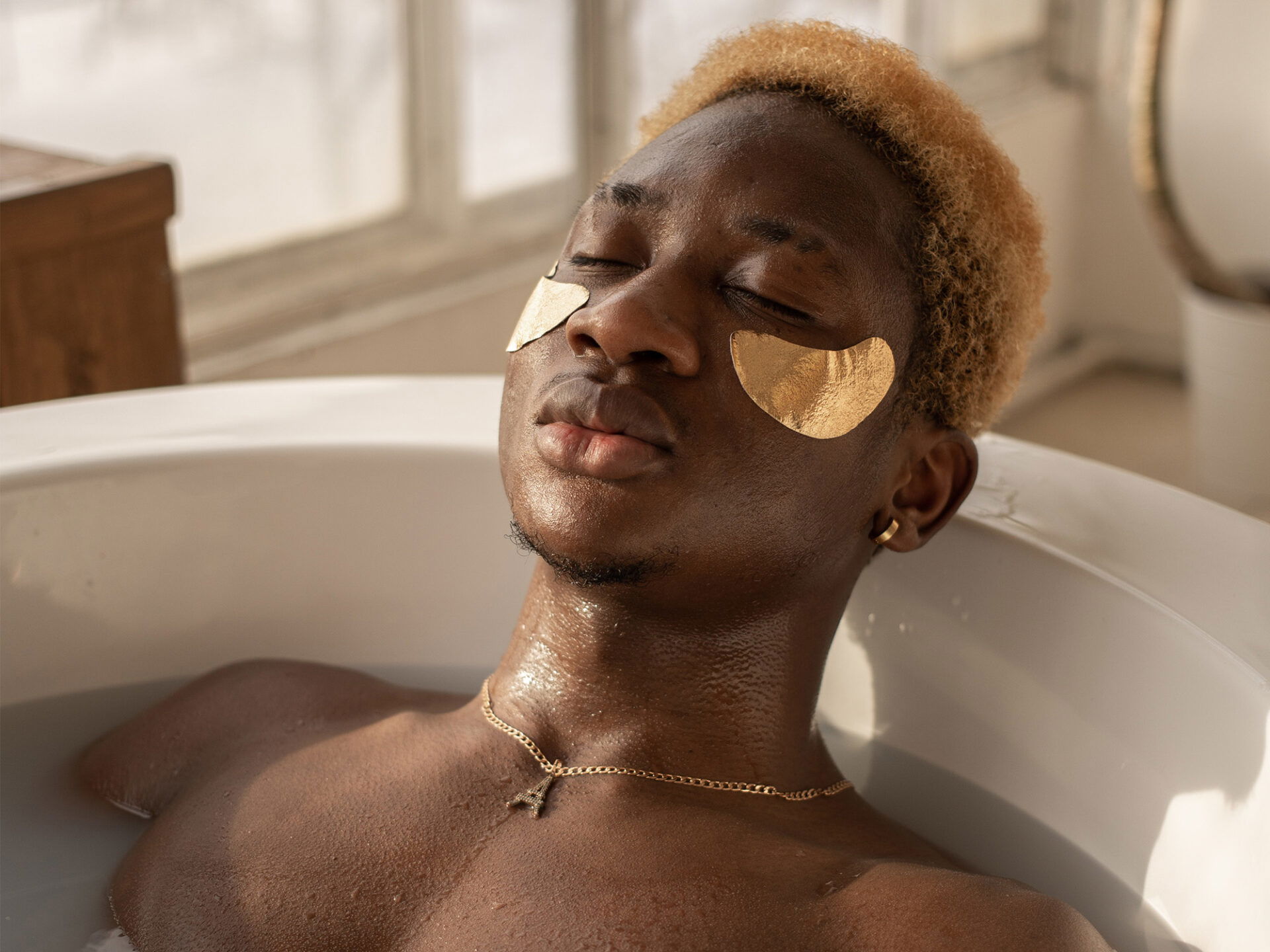 Ein Mann mit geschlossenen Augen und goldfarbenen Augenpflastern liegt in einer Badewanne, umgeben von weichem, natürlichem Licht. Sein Oberkörper ragt aus dem Wasser, während er einen Moment der Erholung und Pflege genießt.