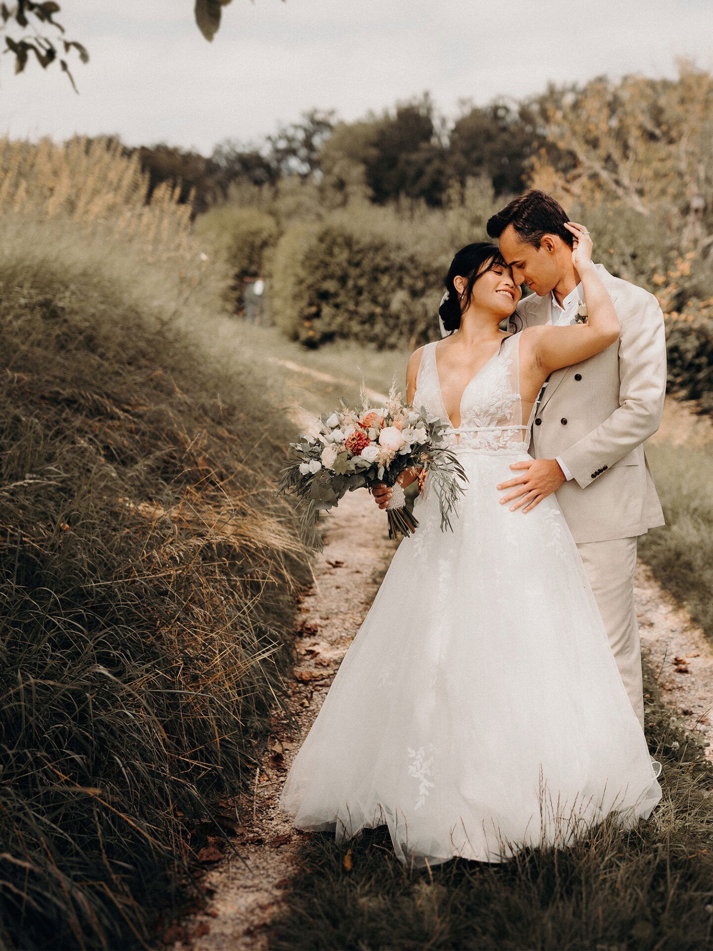 Ein Hochzeitspaar steht liebevoll zusammen auf einem Weg inmitten der Natur. Die Braut, in einem eleganten, weißen Kleid, hält einen üppigen Blumenstrauß, während der Bräutigam in einem beigefarbenen Anzug sie sanft umarmt.