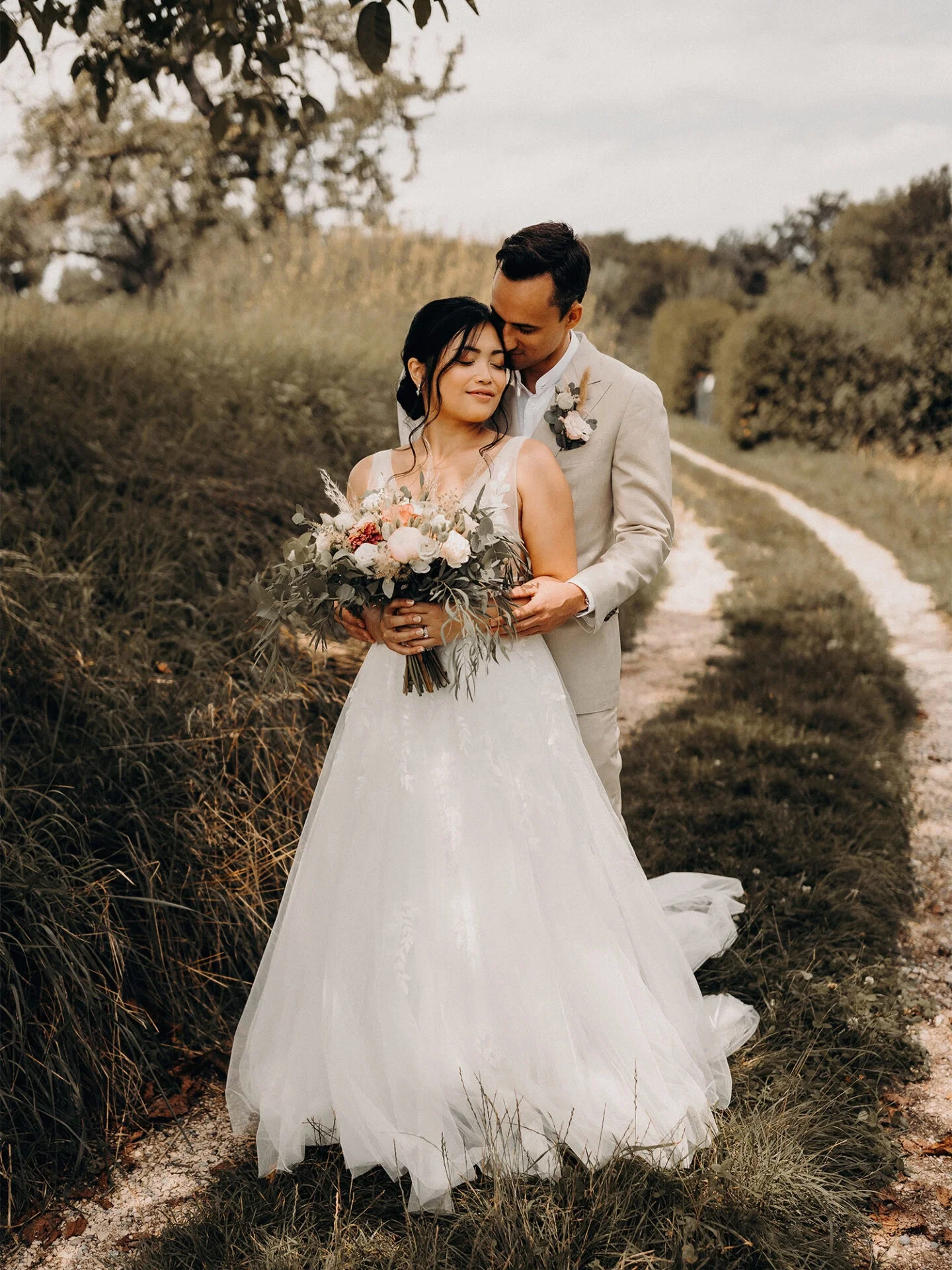 Der Bräutigam küsst seine Braut zärtlich auf die Stirn, während er sie von hinten umarmt. Die Braut trägt ein wunderschönes Kleid und einen Blumenstrauß aus zarten Blüten. Die Szene ist romantisch, mit Bäumen und Gras, die den Weg säumen.