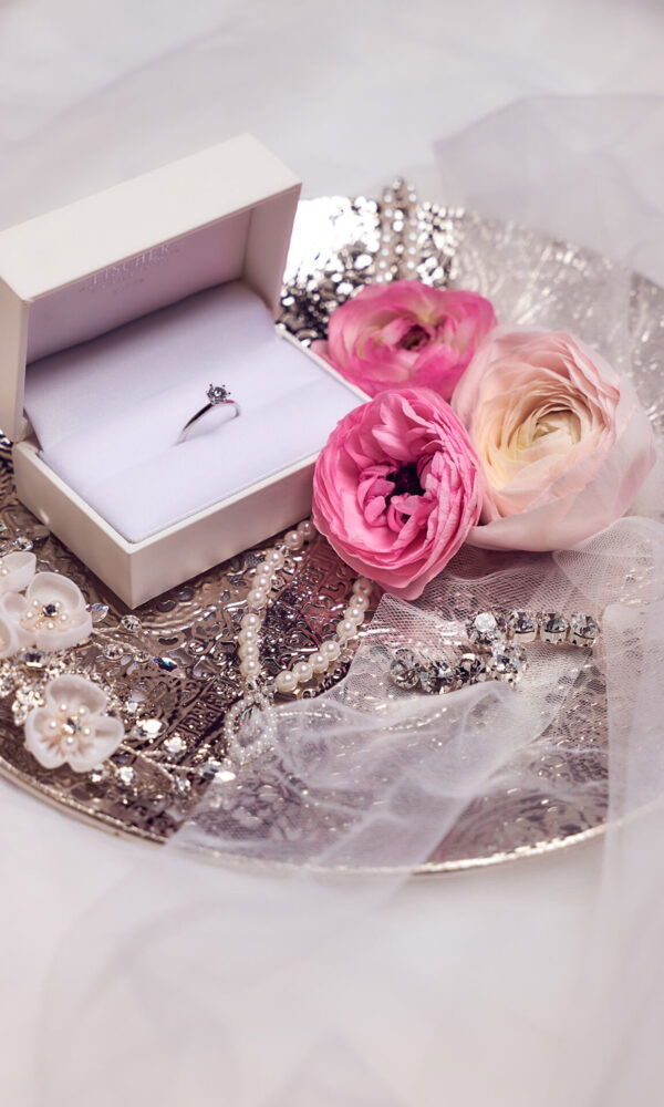 Ein eleganter Verlobungsring in einer weißen Schmuckschatulle liegt auf einem silbernen Tablett. Umgeben wird die Schatulle von rosa Ranunkeln, Perlenketten und funkelndem Brautschmuck. Ein zarter Schleier legt sich über das Arrangement, das eine romantische Atmosphäre schafft.