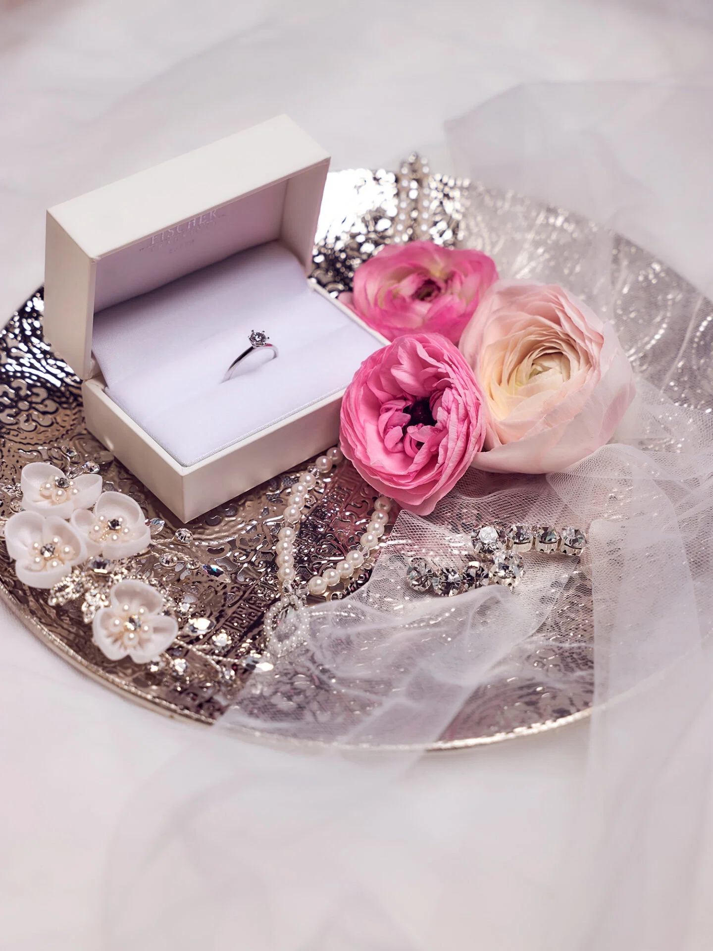 Ein eleganter Verlobungsring in einer weißen Schmuckschatulle liegt auf einem silbernen Tablett. Umgeben wird die Schatulle von rosa Ranunkeln, Perlenketten und funkelndem Brautschmuck. Ein zarter Schleier legt sich über das Arrangement, das eine romantische Atmosphäre schafft.