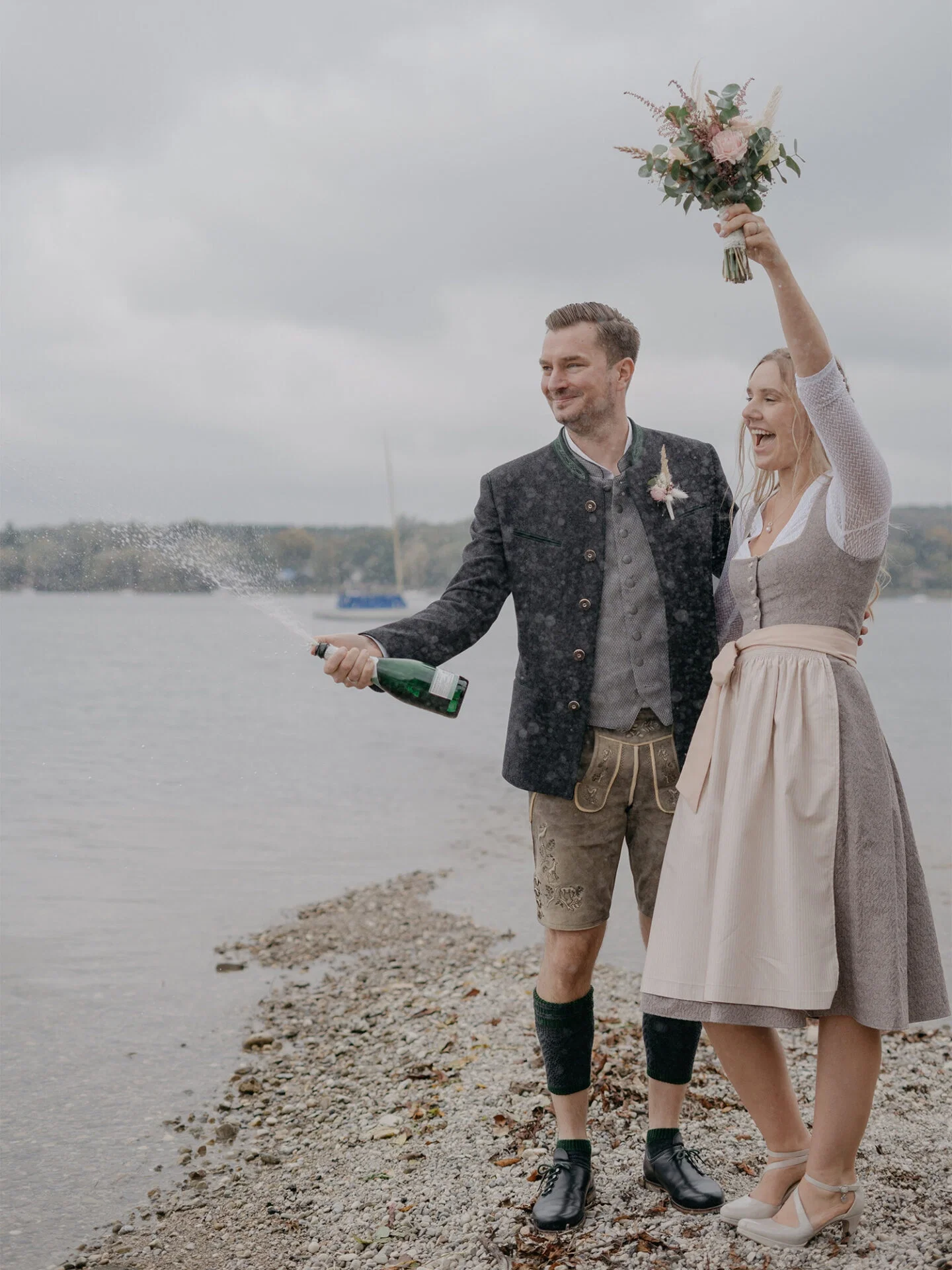 Ein Paar in traditioneller bayerischer Tracht steht an einem Seeufer. Der Bräutigam spritzt freudig mit einer Champagnerflasche, während die Braut strahlend einen Blumenstrauß in die Luft hält.