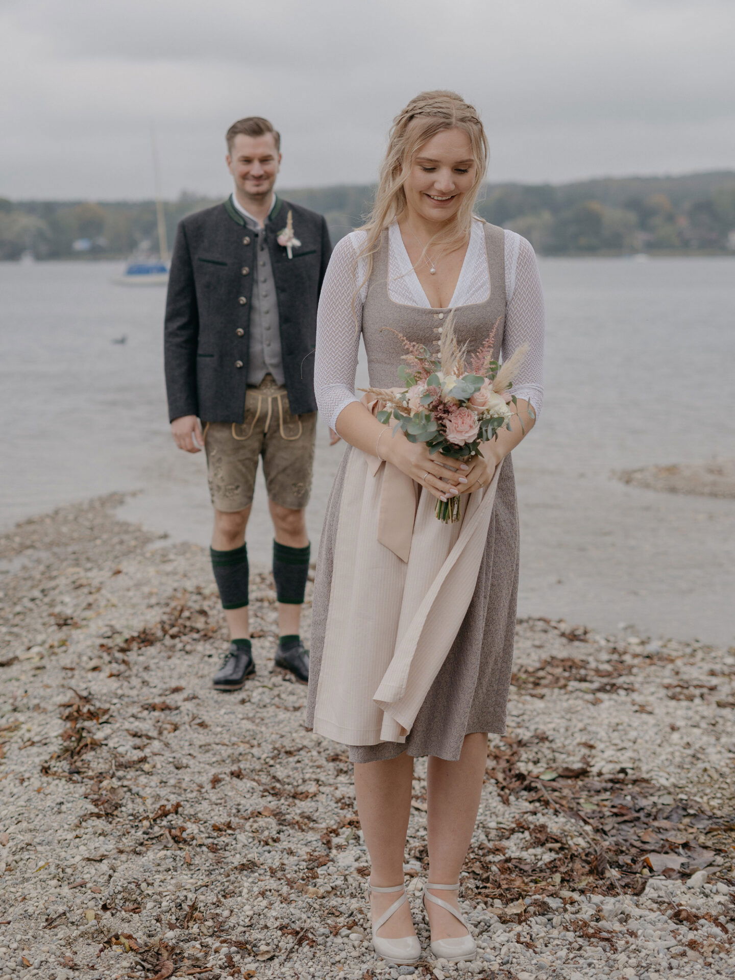 Die Braut, in einem Dirndl, steht lächelnd am Seeufer und hält einen Blumenstrauß. Im Hintergrund steht der Bräutigam in Lederhosen und einem Trachtenjanker.