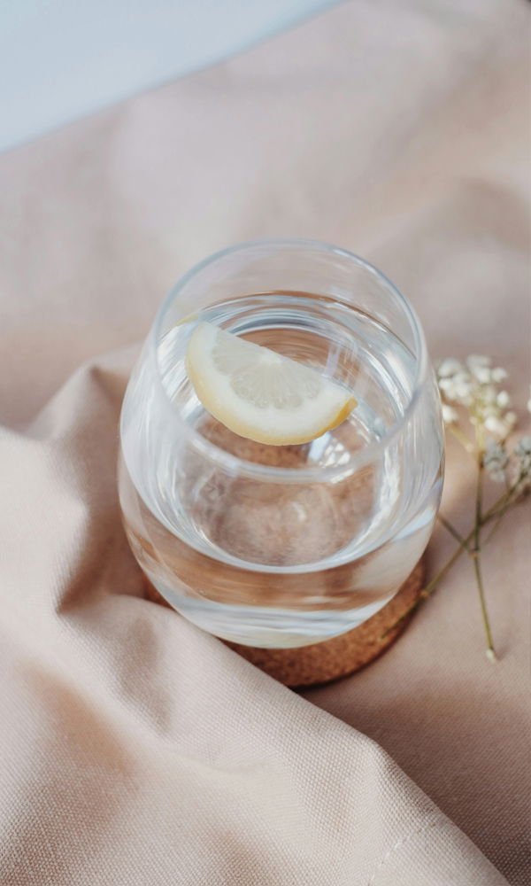 Nahaufnahme eines Wasserglases mit einer Zitronenscheibe. Das Glas steht auf einem weichen beigen Untergrund, dekoriert mit minimalistischen Details.