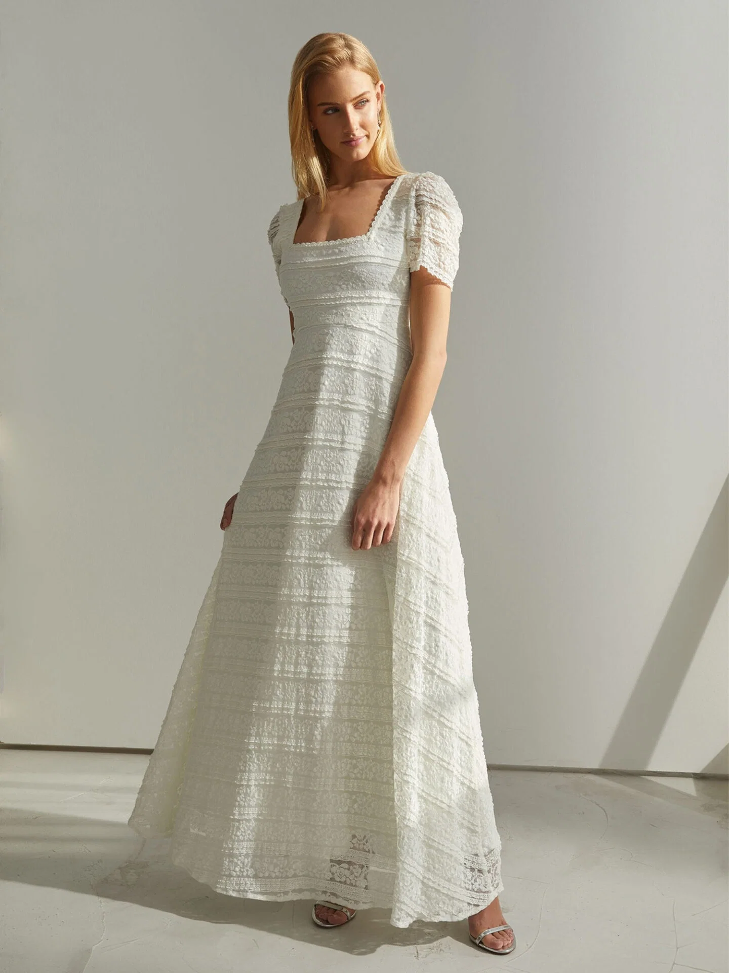 Eine Frau steht in einem hellen Raum und trägt ein schlichtes Empire-Kleid mit kurzen Ärmeln und einem quadratischen Ausschnitt. Ihr Kleid wirkt leicht und strukturiert, während sie ruhig und selbstbewusst in die Ferne blickt.