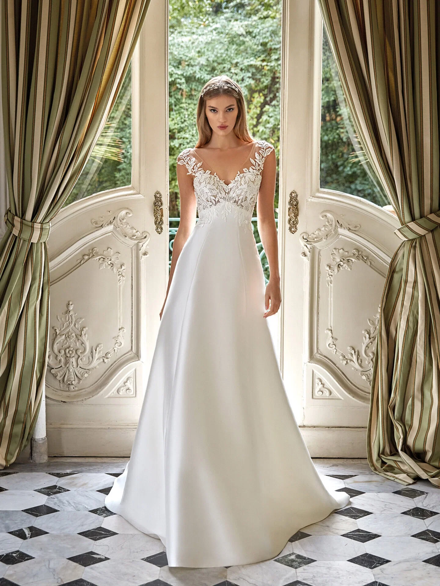 Eine Braut posiert in einem eleganten, klassischen Raum in einem weißen Empire-Kleid mit floralen Spitzenakzenten. Hinter ihr öffnen sich große Türen, die den Blick auf einen üppigen Garten freigeben.