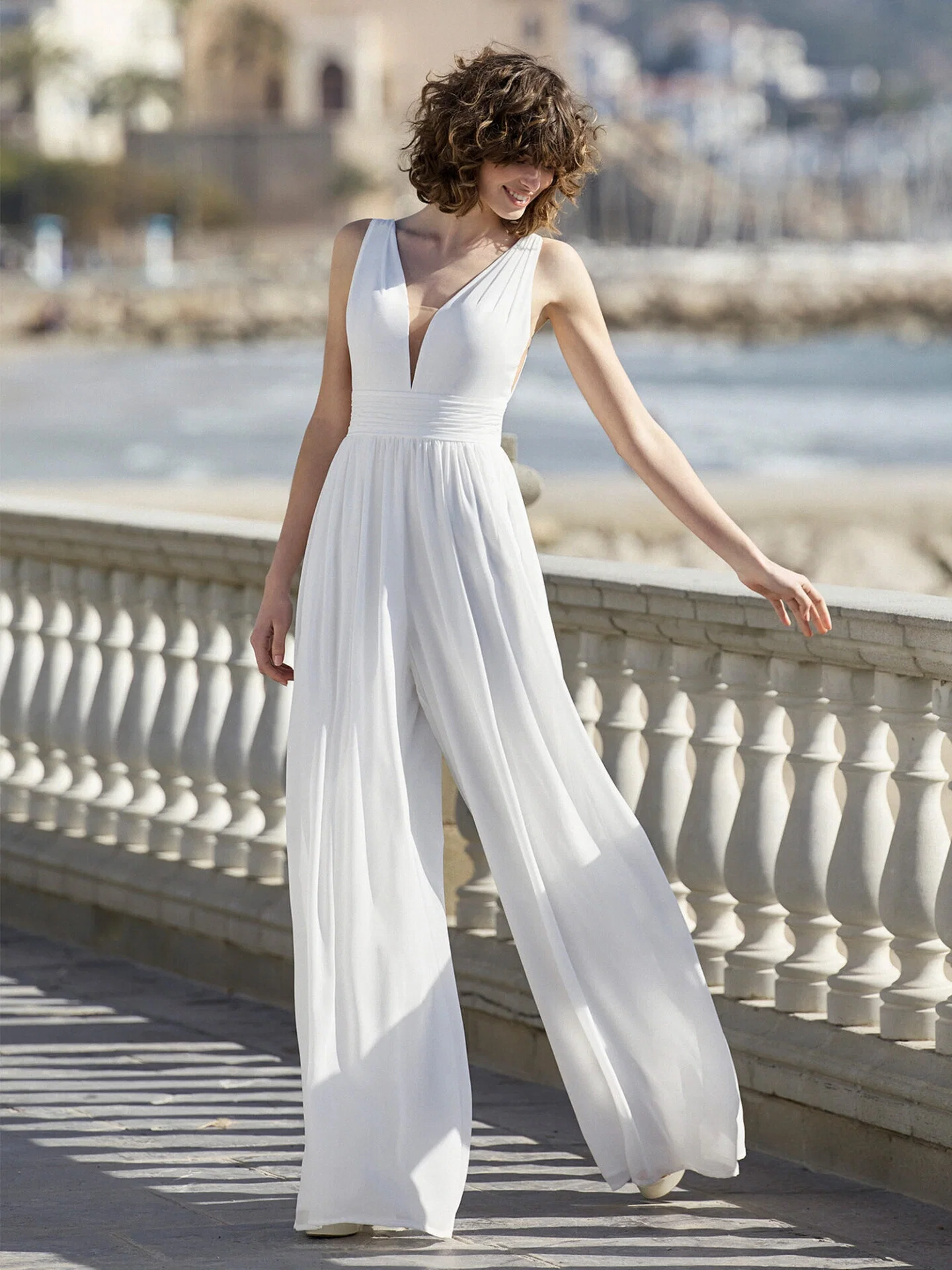 Eine Braut trägt einen modernen, weiten Jumpsuit mit tiefem V-Ausschnitt und posiert an einer Promenade mit Blick auf das Meer. Der luftige Schnitt des Outfits verleiht dem Look eine lockere, sommerliche Eleganz.