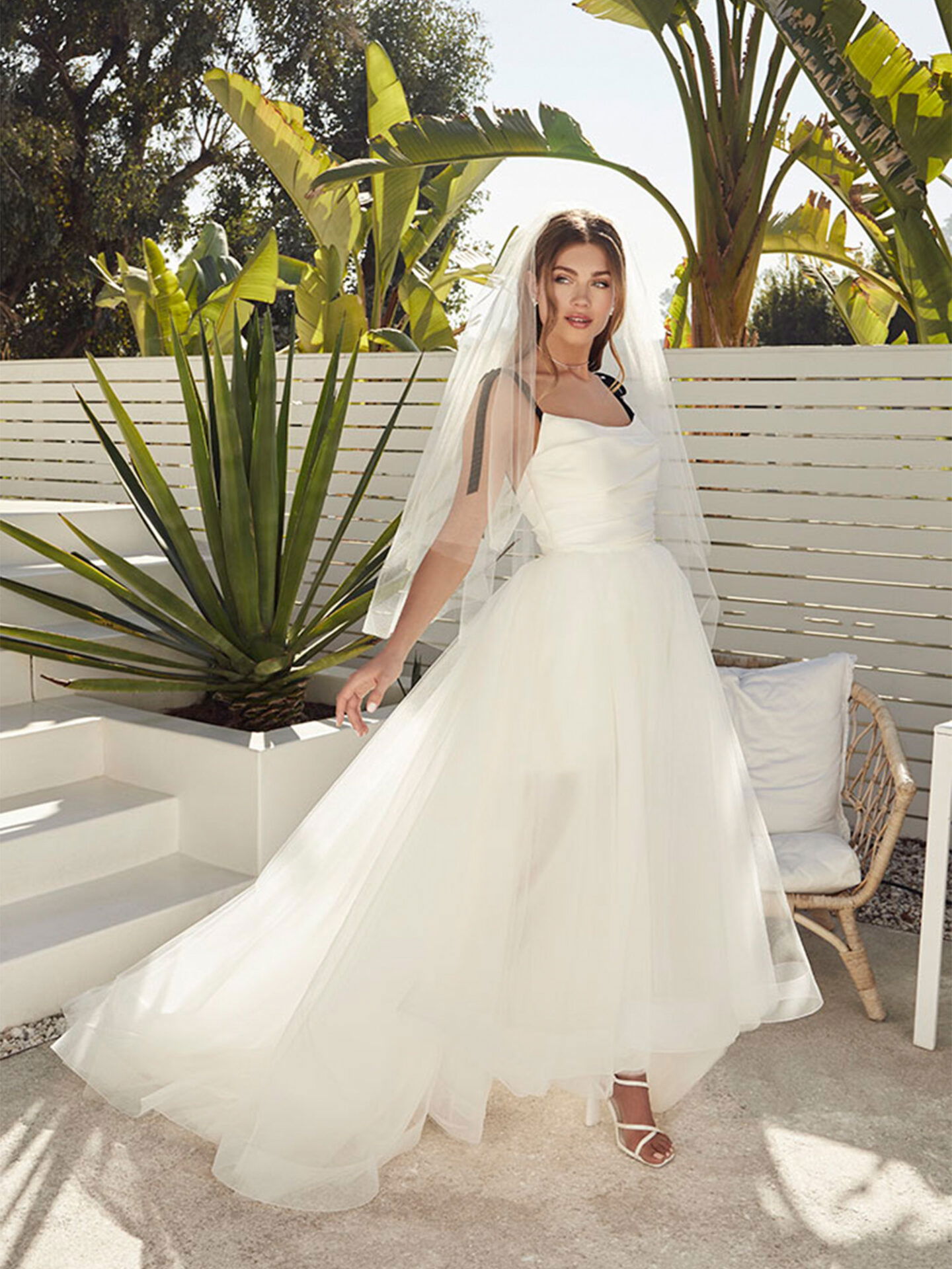 Eine Braut in einem weißen Tüllkleid steht vor einer weißen Wand und tropischen Pflanzen. Sie trägt einen langen, transparenten Schleier, der im Sonnenlicht glänzt, während der voluminöse Rock ihres Kleides in der Brise weht.