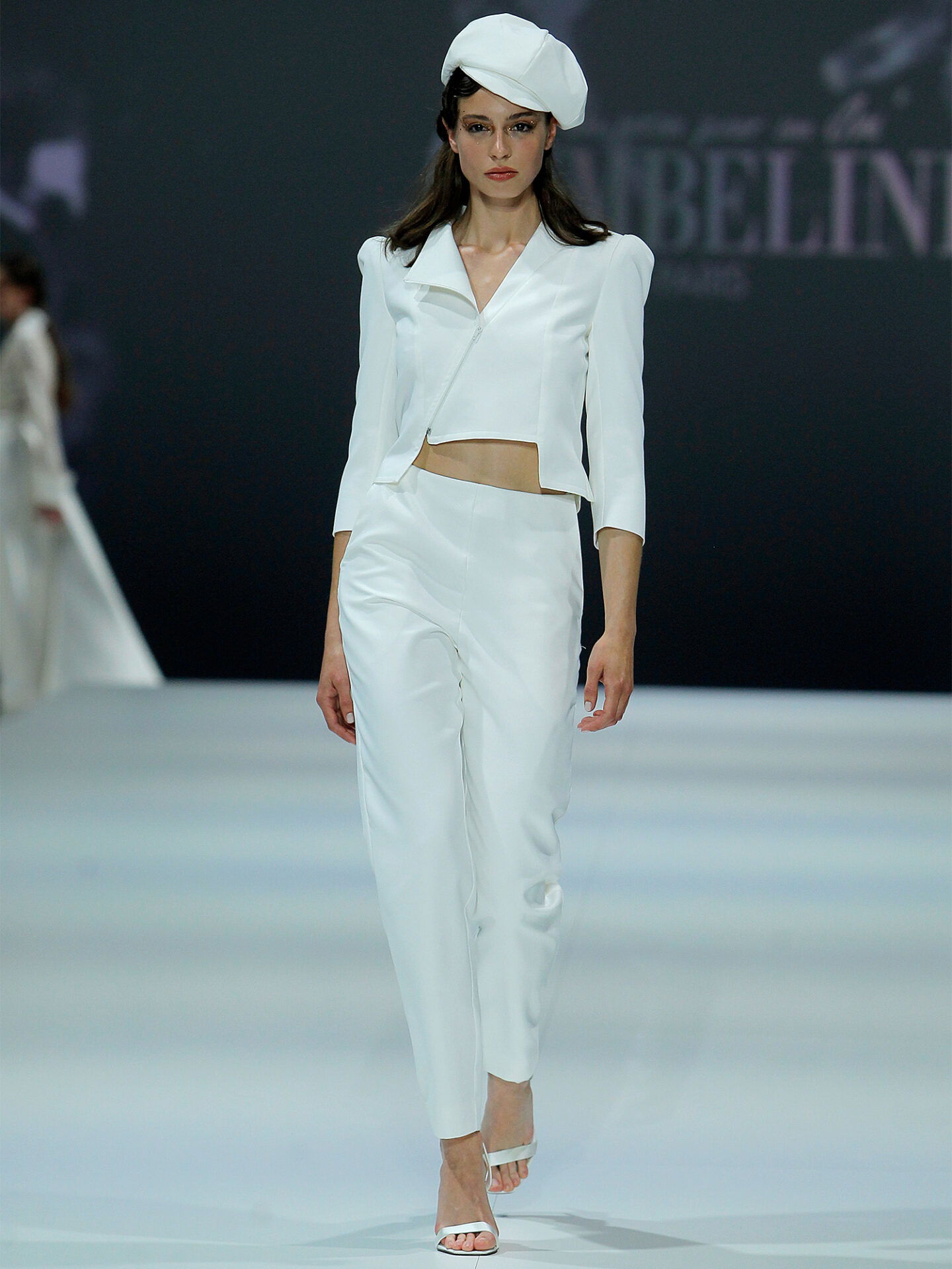 Eine Frau läuft über den Laufsteg in einem zweiteiligen Brautoutfit, bestehend aus einem kurzen Blazer und einer weißen Hose. Der schlichte und elegante Look wird durch eine weiße Mütze ergänzt, was dem Outfit eine besondere Note verleiht.