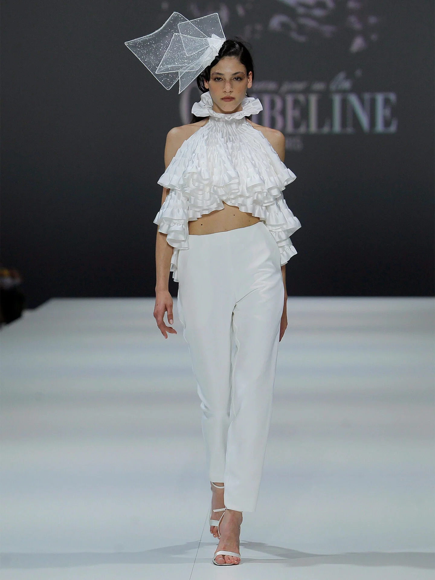 Ein Model präsentiert auf dem Laufsteg einen modischen Zweiteiler mit einem voluminösen, rüschenbesetzten Oberteil und einer schlichten Hose. Der auffällige Kopfschmuck in geometrischem Design rundet den extravaganten Look ab.