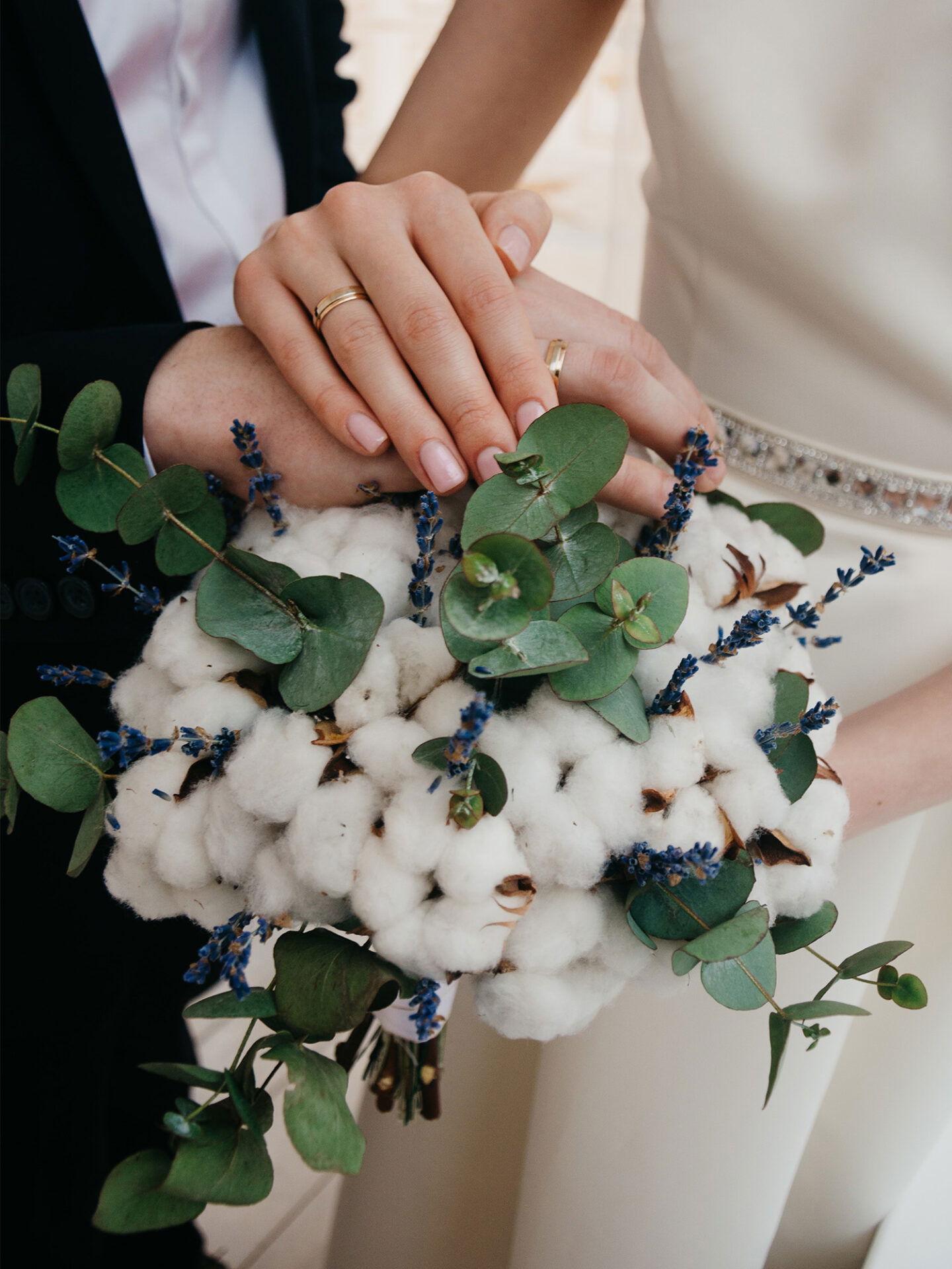 Die Hände eines frisch verheirateten Paares, die symbolisch über einem einzigartigen Strauß aus Baumwollblüten und Lavendel liegen.