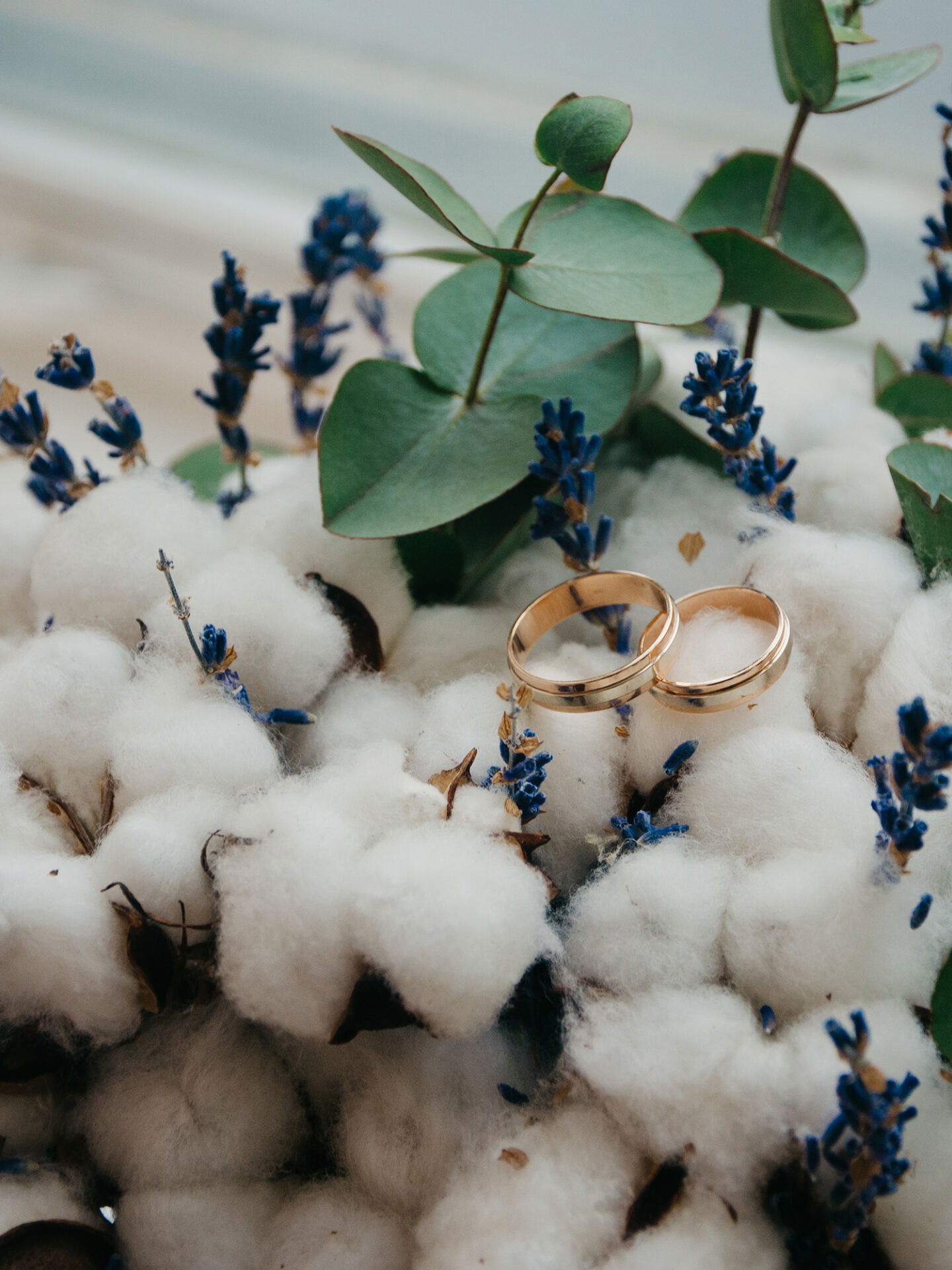 Detailfoto eines Brautstraußes aus Baumwolle, Lavendel und Eukalyptus, auf dem zwei schlichte goldene Trauringe liegen.