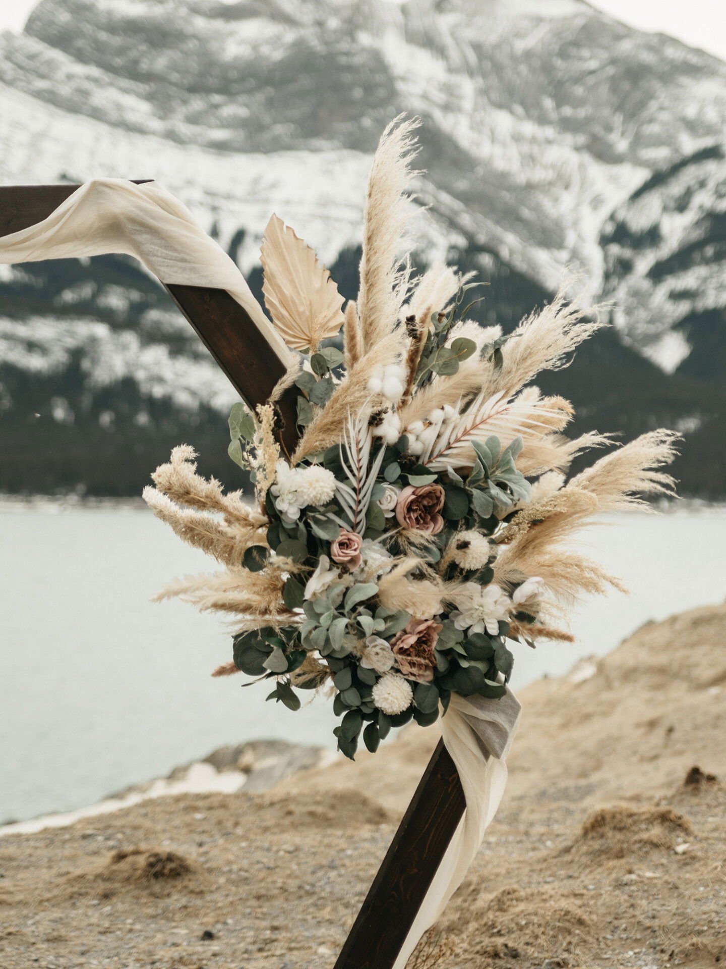Rustikaler Blumenstrauß aus getrockneten Pampasgräsern und Eukalyptus, der als Teil einer Hochzeitsdekoration vor schneebedeckten Bergen präsentiert wird.