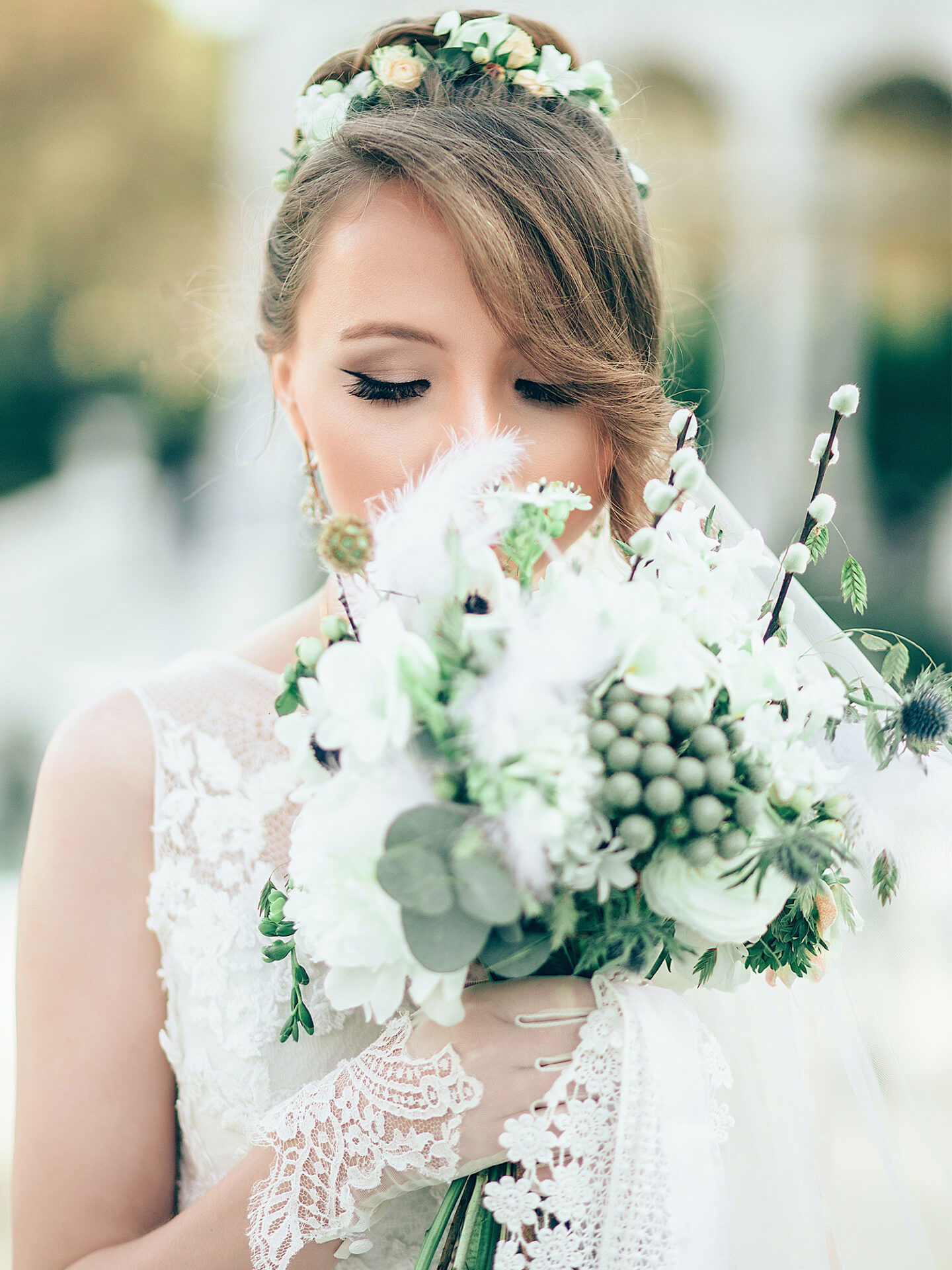 Nahaufnahme einer Braut mit einem Blumenstrauß aus weißen Blüten, Federn und grünen Zweigen, die liebevoll die Komposition betrachtet.