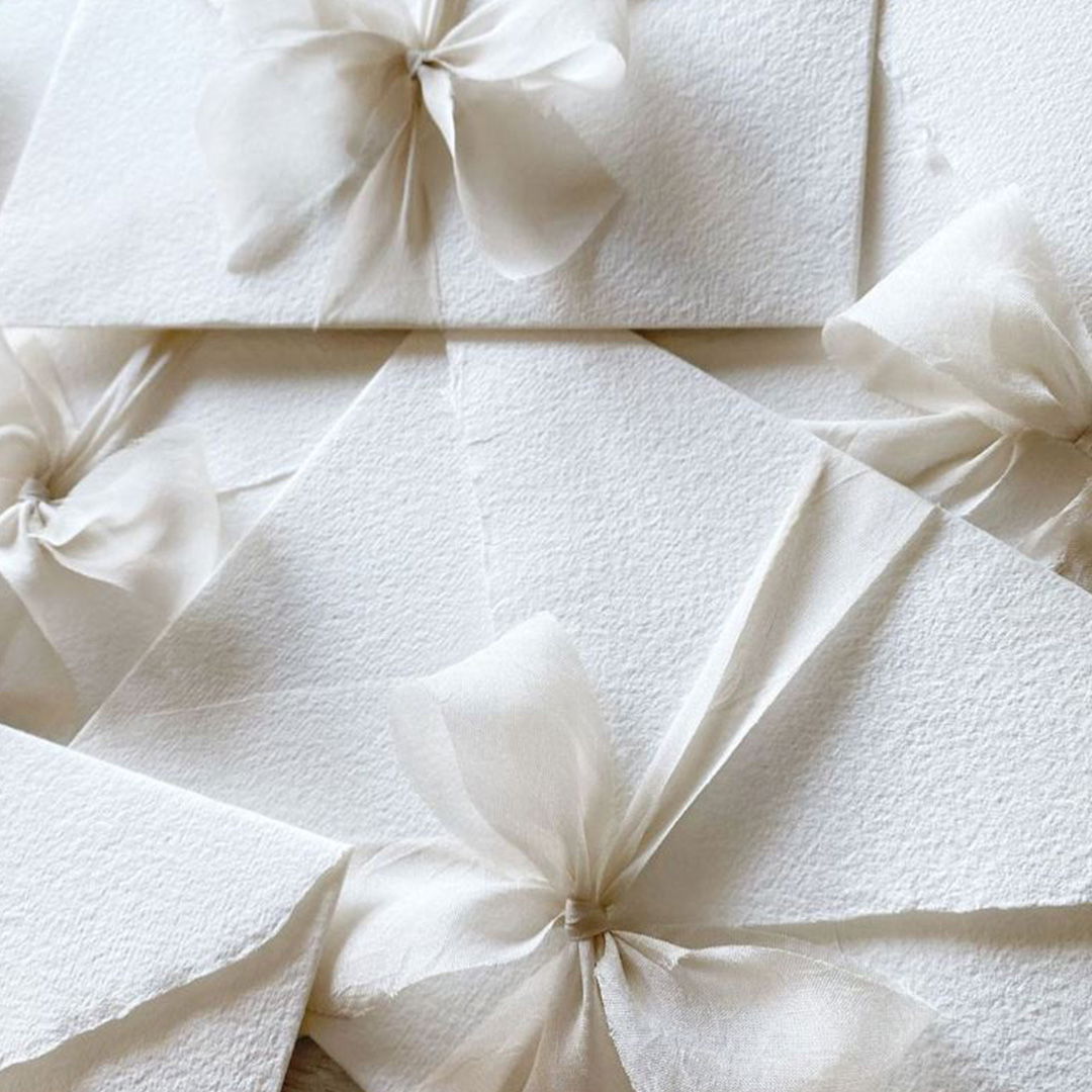 Ein Stapel eleganter, weißer Umschläge, die mit filigranen, durchsichtigen Schleifen geschmückt sind. Die zarten Schleifen verleihen den Umschlägen eine festliche, handgemachte Note, passend für eine besondere Feier.