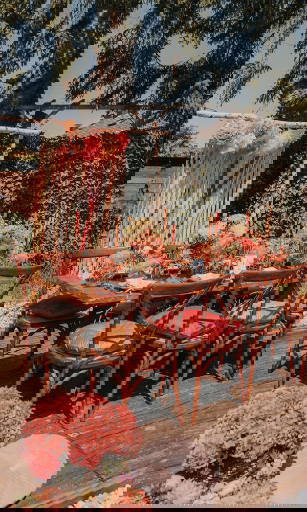 Rustikale Hochzeitstafel unter freiem Himmel, dekoriert mit floralen Arrangements, Holzdetails und farbigen Stuhlkissen in einem grünen Garten.