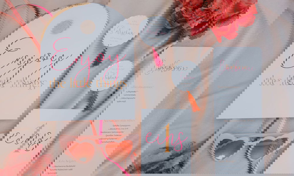 Kreatives Hochzeitspapeterie-Set auf beigem Untergrund mit liebevoll gestalteten Karten, goldenen Akzenten, farbigen Quasten und einer rosa Sonnenbrille in Herzform, eingerahmt von Blumen.