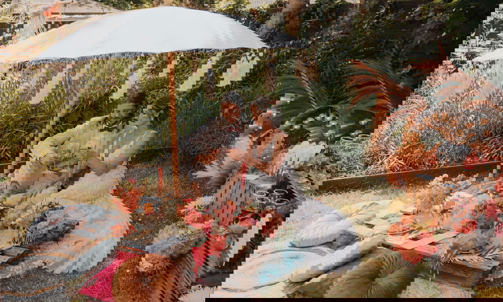 Rustikale Gartenhochzeit mit einem Hochzeitspaar bei einem Picknick unter einem geschmückten Sonnenschirm, umrahmt von Boho-Dekoration und leuchtenden Blumen.