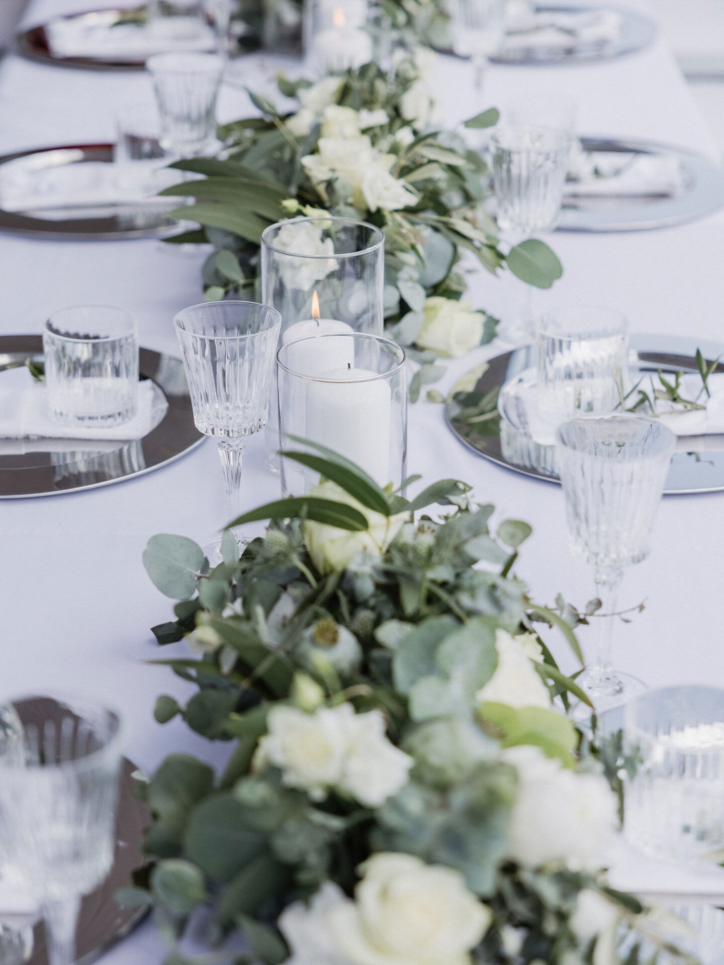 Ein gedeckter Hochzeitstisch mit kristallklaren Gläsern, dezenten weißen Kerzen und einer floralen Dekoration aus Eukalyptus und Rosen. Der Brautstrauß, der aus Eukalyptus und weißen Blumen besteht, wird von der Braut in einem offenen Feld getragen.
