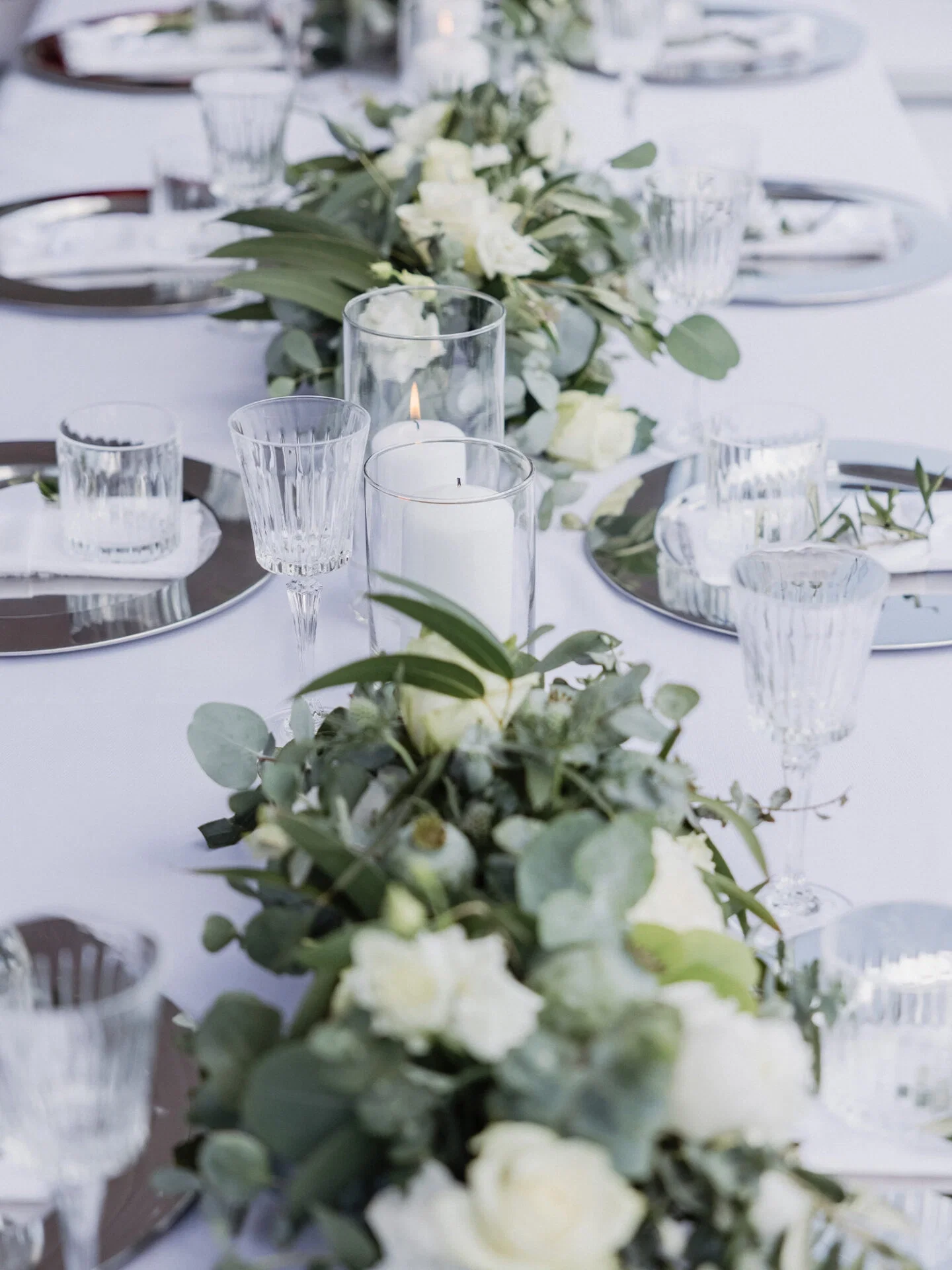 Ein gedeckter Hochzeitstisch mit kristallklaren Gläsern, dezenten weißen Kerzen und einer floralen Dekoration aus Eukalyptus und Rosen. Der Brautstrauß, der aus Eukalyptus und weißen Blumen besteht, wird von der Braut in einem offenen Feld getragen.