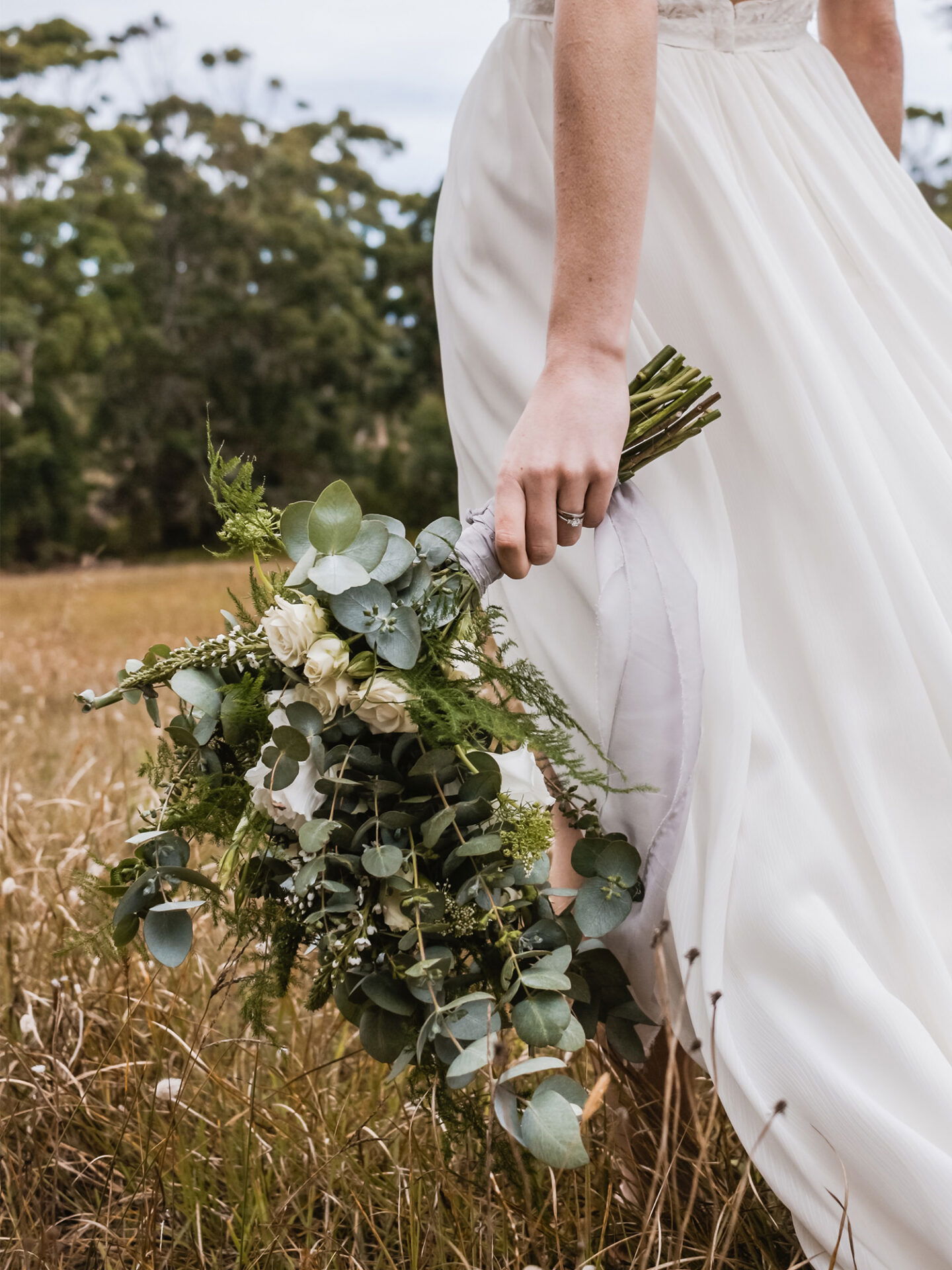 Die Braut hält einen natürlichen, grünen Hochzeitsstrauß mit Eukalyptus und weißen Blumen, während sie in einem weitläufigen Feld spazieren geht.