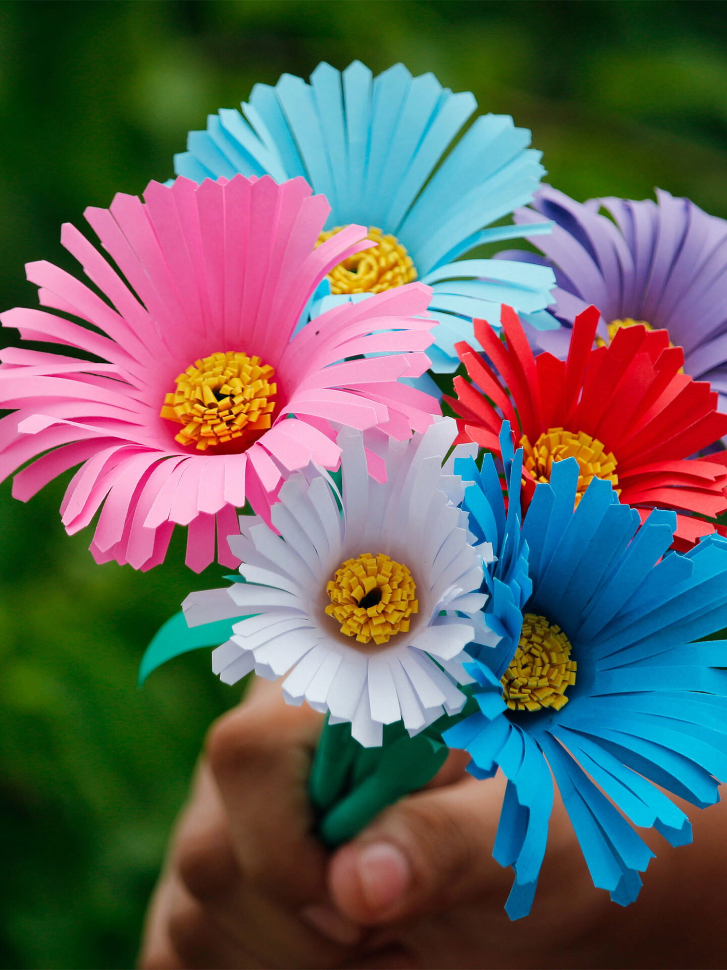 Lebendig und fröhlich: Ein Strauß aus farbenfrohen, sorgfältig geschnittenen Papierblumen in Pink, Blau, Rot und Weiß, präsentiert in einer Hand.