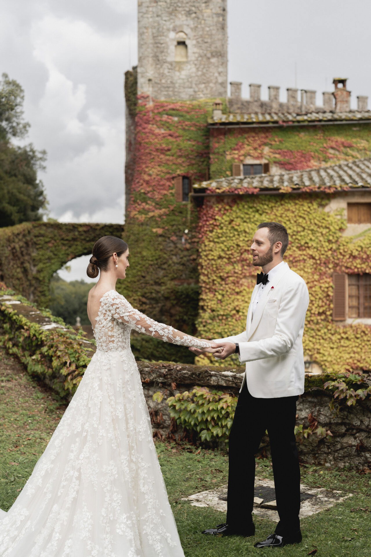 Hochzeitspaar, das sich vor einer mit Efeu überwucherten Steinmauer gegenübersteht und sich die Hände reicht. Die Braut trägt ein kunstvolles Spitzenkleid mit langen Ärmeln, der Bräutigam einen klassischen weißen Smoking.