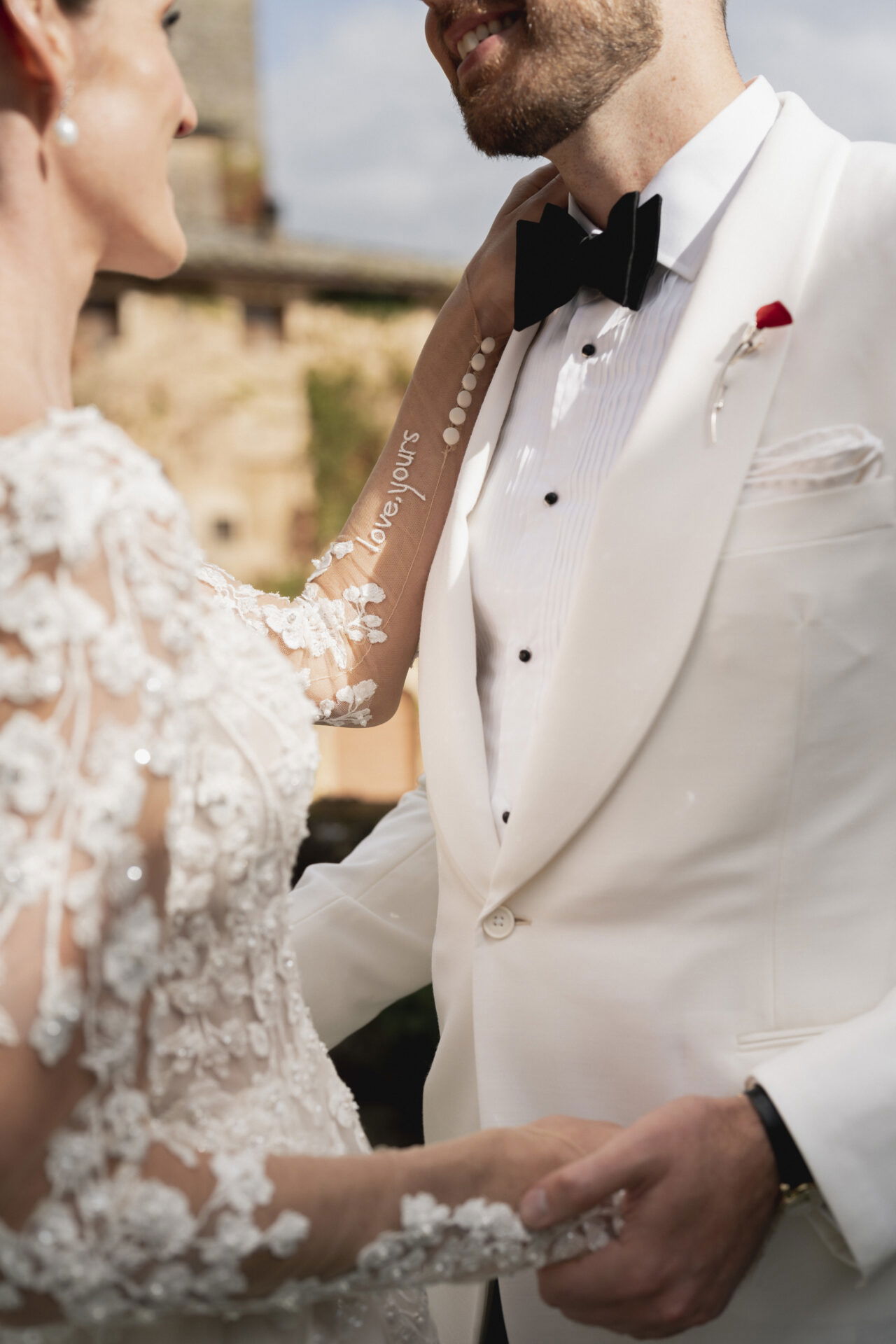Romantischer Moment zwischen Braut und Bräutigam in eleganter Hochzeitskleidung. Die Braut trägt ein florales Spitzenkleid, während der Bräutigam einen weißen Smoking mit rotem Ansteckblumen trägt. Auf dem Ärmel der Braut ist 'Love, yours' eingestickt.