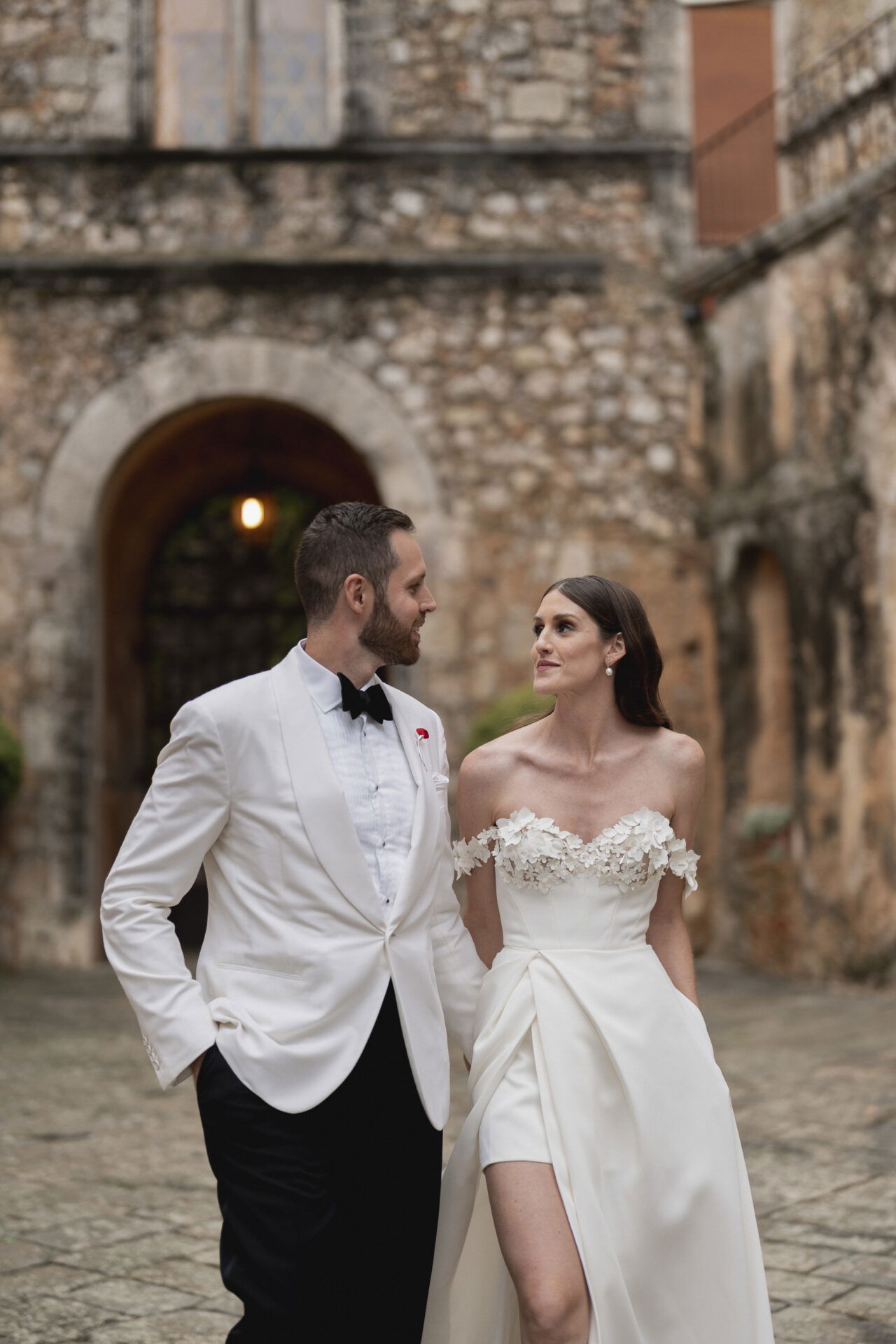 Braut und Bräutigam blicken sich liebevoll an, während sie durch einen alten, charmanten Innenhof spazieren. Die Braut trägt ein schulterfreies Kleid mit hohem Schlitz, der Bräutigam einen klassischen weißen Smoking.