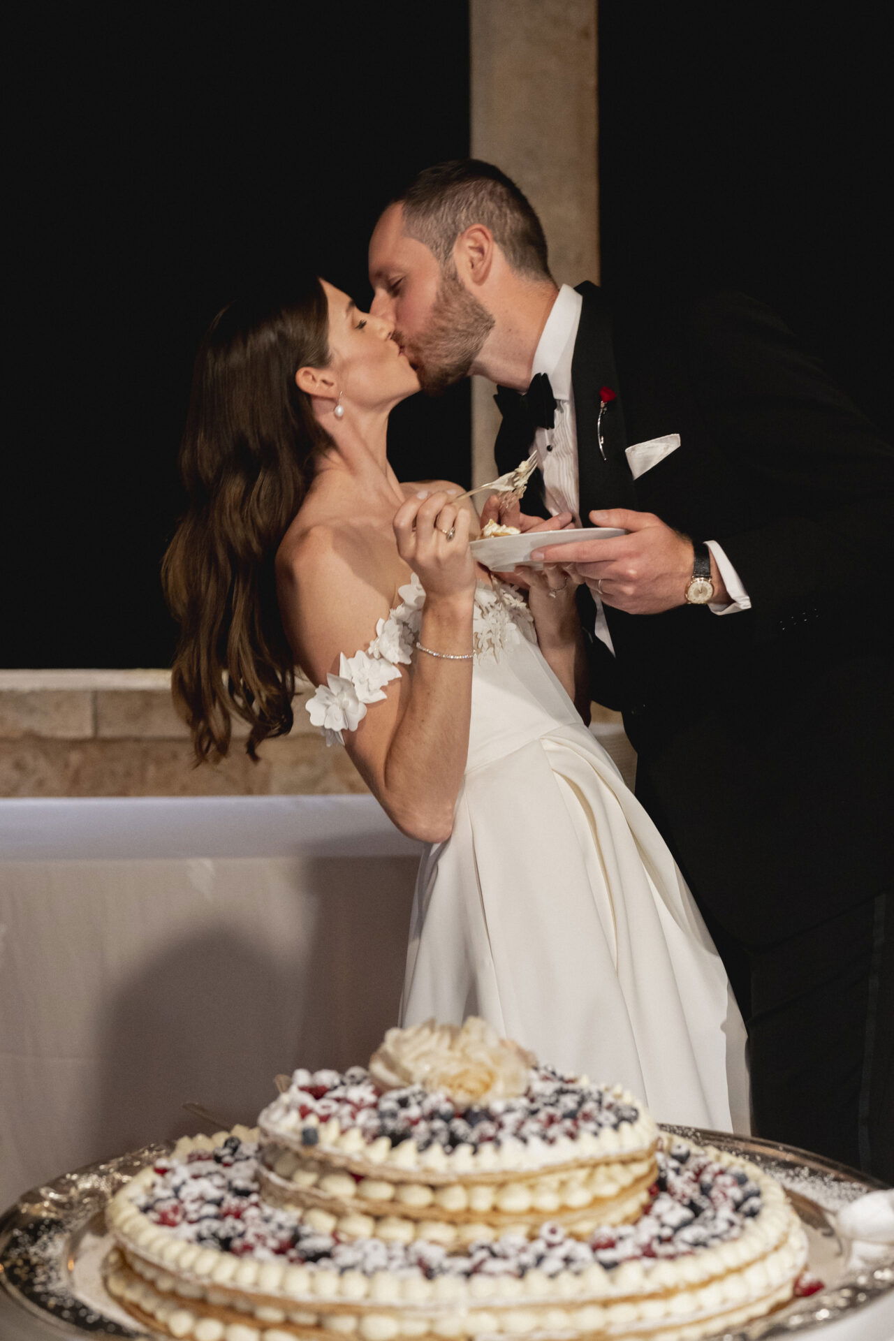 Der Moment des Hochzeitstorten-Anschnitts wird von einem Kuss zwischen Braut und Bräutigam gekrönt. Beide halten Teller mit Kuchen in der Hand, während die kunstvoll verzierte Torte mit frischen Beeren im Vordergrund steht.