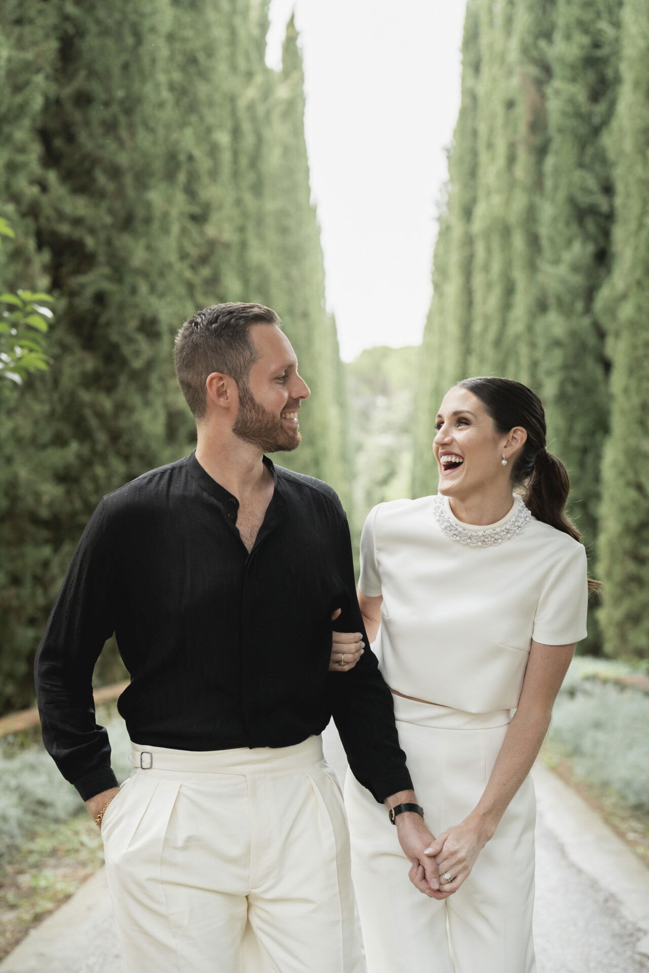 Ein Paar genießt einen Spaziergang in einer von Bäumen umgebenen Allee. Beide lachen glücklich miteinander, der Mann trägt Schwarz und Weiß, die Frau trägt ein minimalistisches, weißes Outfit und wirkt fröhlich und entspannt.