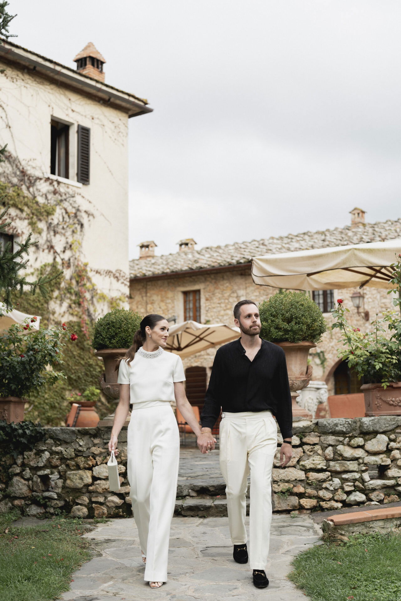 Ein lächelndes Paar geht Hand in Hand durch die Gärten eines historischen Anwesens, umgeben von Natur und Steinmauern. Beide sind in kontrastierenden Outfits gekleidet: Die Frau in Weiß, der Mann in Schwarz und Weiß.