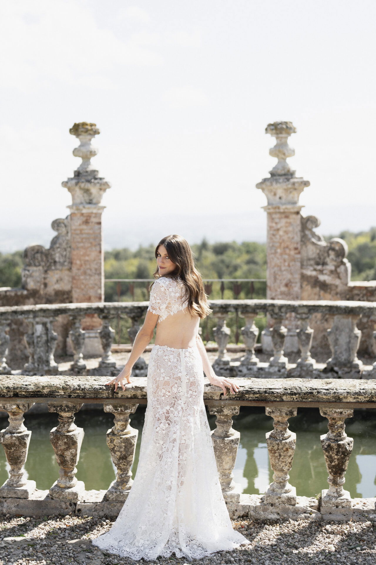 Eine Braut posiert in einem transparenten Spitzenkleid mit offenem Rücken auf einer antiken Terrasse, umgeben von steinernen Säulen. Sie schaut zurück in die Kamera und strahlt eine ruhige Eleganz aus.