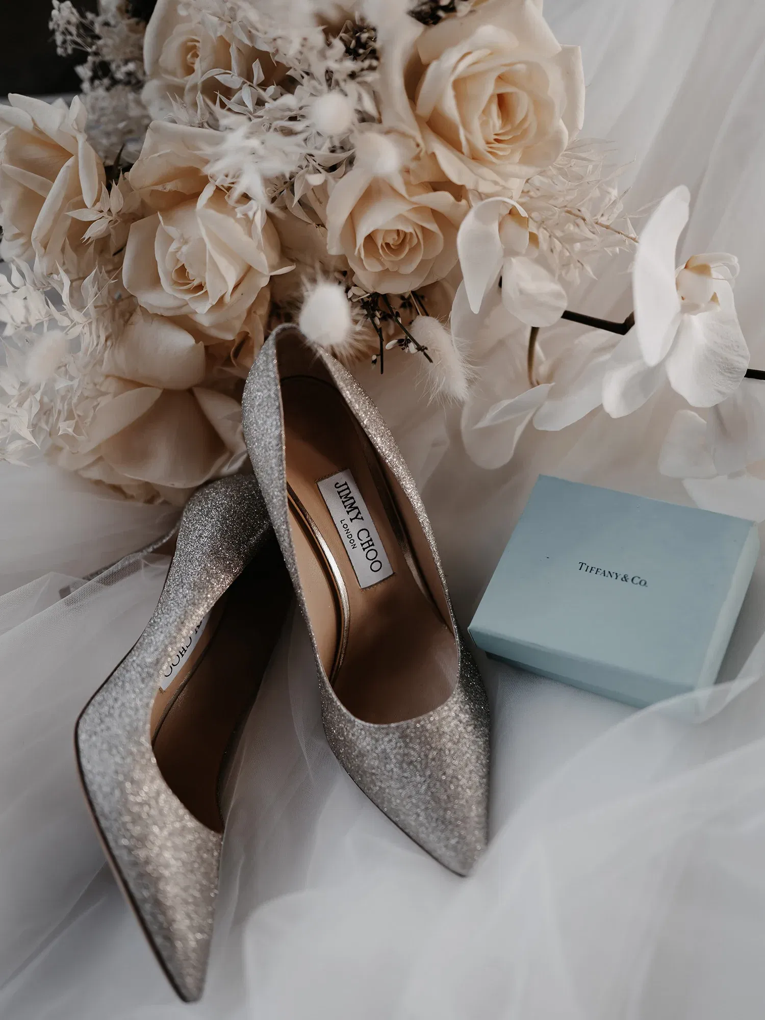 Glitzernde Pumps von Jimmy Choo, begleitet von einem Brautstrauß aus hellen Rosen und einer Tiffany & Co. Geschenkbox, sorgfältig auf einem Brautschleier drapiert.