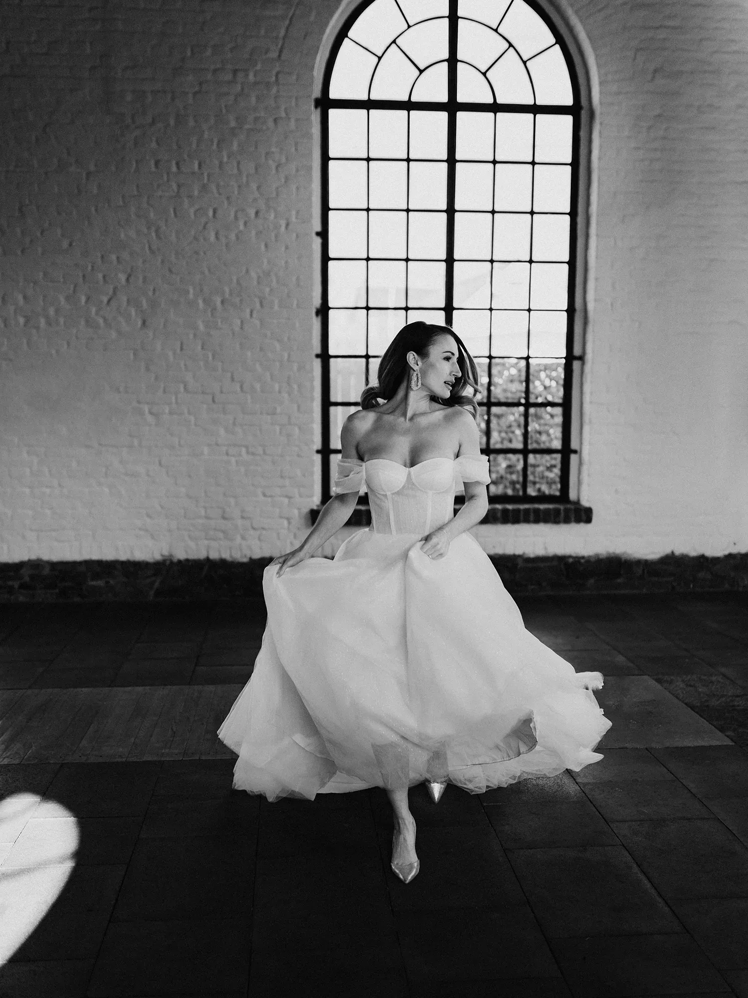 Eine Braut in einem zeitgenössischen Hochzeitskleid wirbelt durch einen Raum mit einer rustikalen Steinwand und einem hohen Bogenfenster.