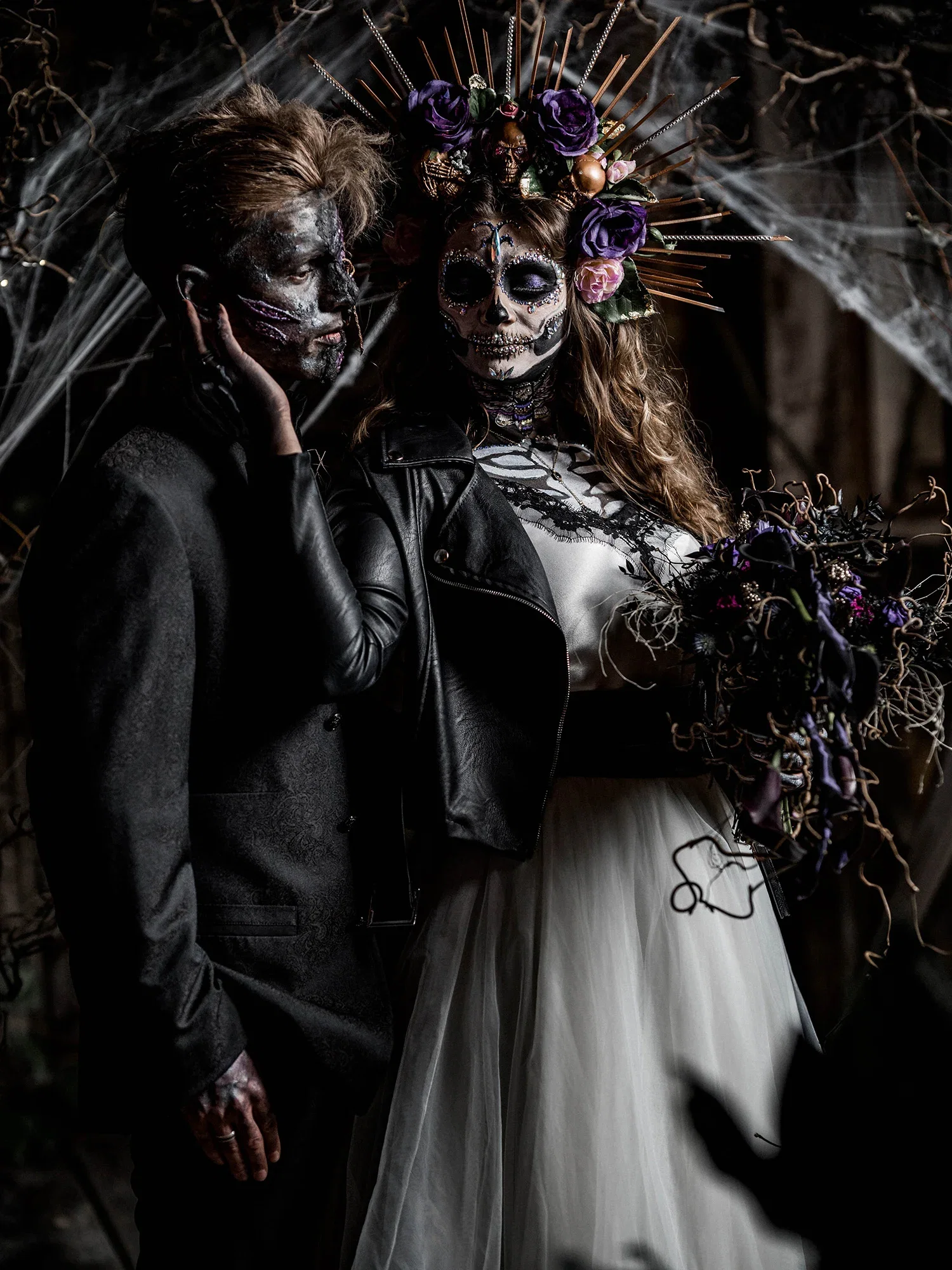Ein Gothic-Brautpaar posiert mit detaillierter Halloween-Schminke und kunstvollen Blumenarrangements in einer düsteren, geheimnisvollen Umgebung.