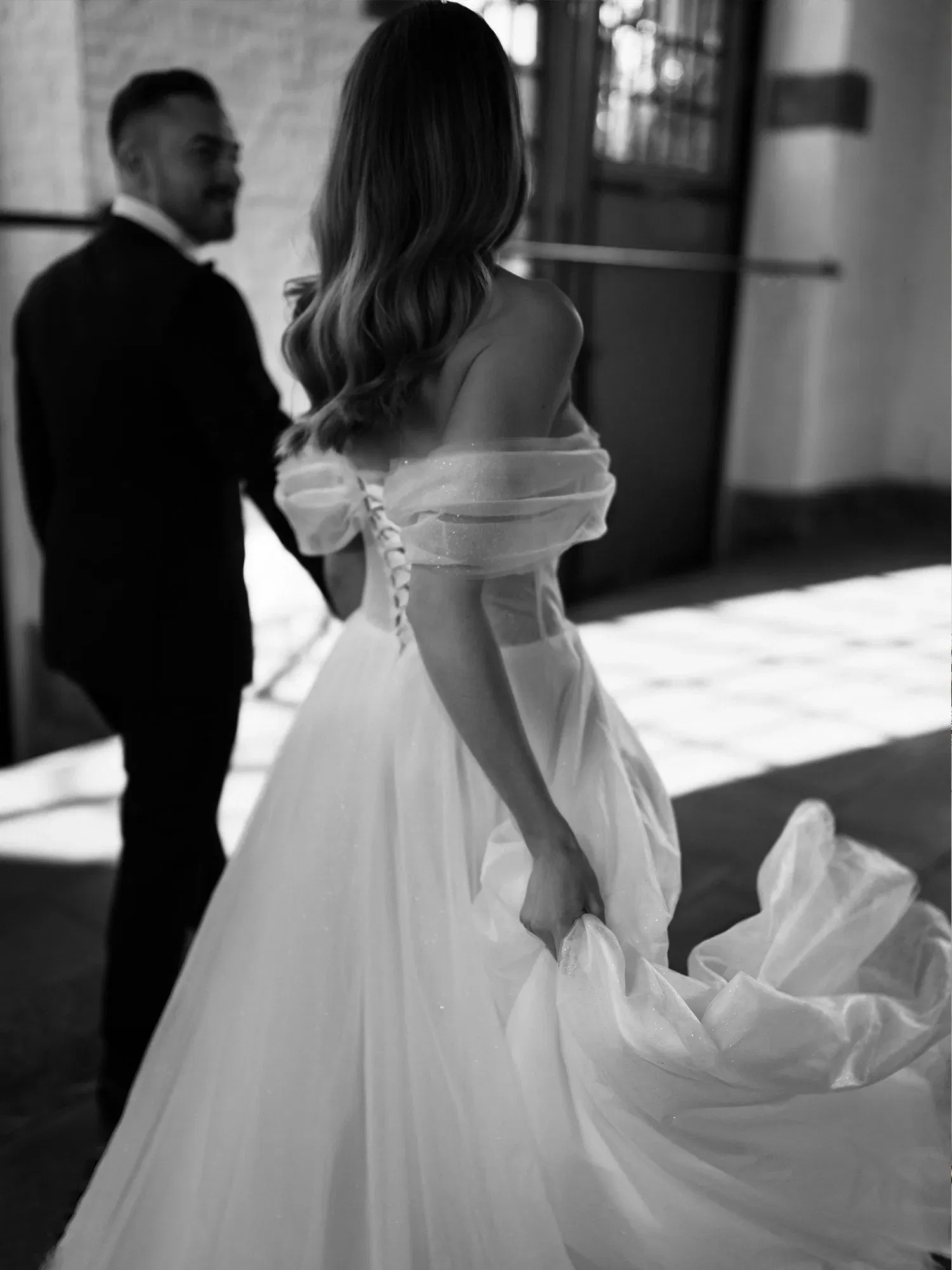 Romantisches Schwarz-Weiß-Bild eines Brautpaares, die Braut in einem Off-Shoulder-Kleid, der Bräutigam schaut lächelnd auf sie, während sie gemeinsam durch einen Saal gehen.
