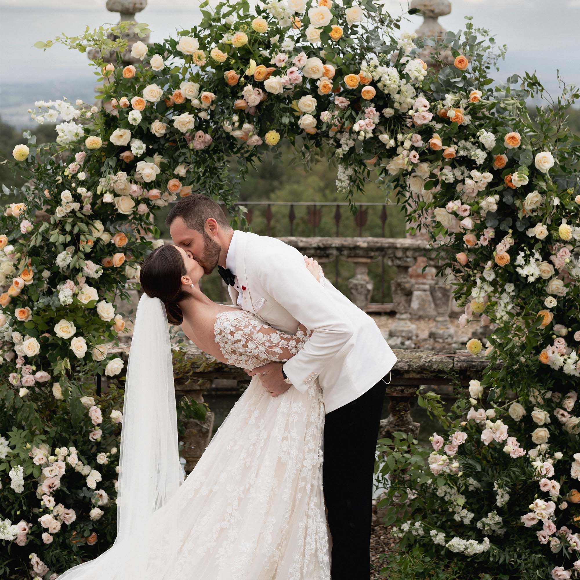 Hochzeitspaar in einem romantischen Moment, umgeben von einem Blumenbogen mit weißen und gelben Rosen. Die Braut trägt ein prächtiges Kleid mit Spitze, während der Bräutigam sie in einem weißen Jackett sanft küsst.