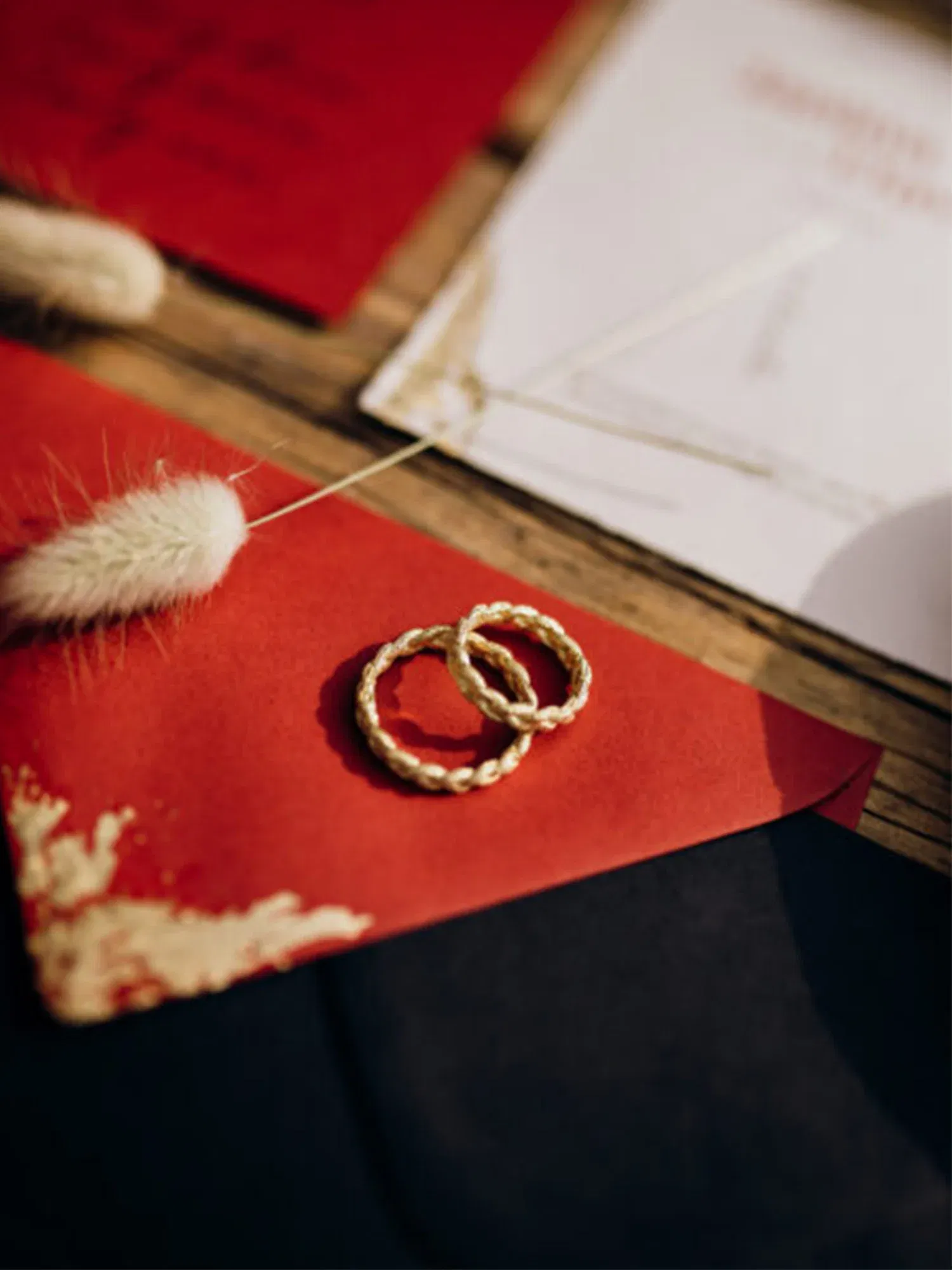 Zwei miteinander verschlungene goldene Eheringe liegen dekorativ auf einem roten Umschlag, begleitet von eleganten, beigen Federn.