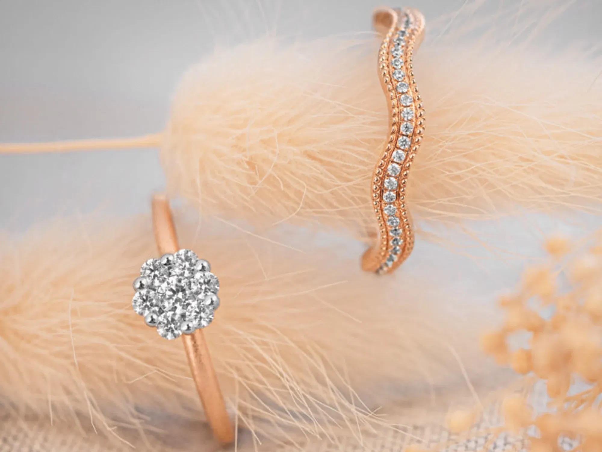 Ein roségoldener Ring mit funkelnden Diamanten in einem wellenförmigen Design liegt neben einem Ring mit einem runden Diamanten, der von mehreren kleinen Steinen eingefasst ist, vor einem sanften, beigen Hintergrund.