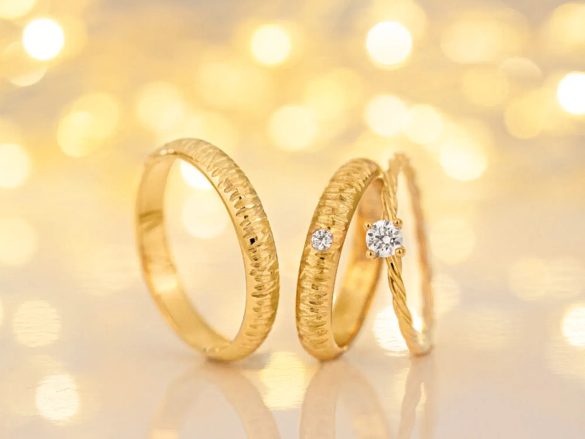 Drei feine, goldene Ringe, jeder mit einem einzelnen funkelnden Diamanten verziert, stehen vor einem sanften, goldenen Bokeh-Hintergrund, der die Eleganz und Raffinesse der Schmuckstücke unterstreicht.