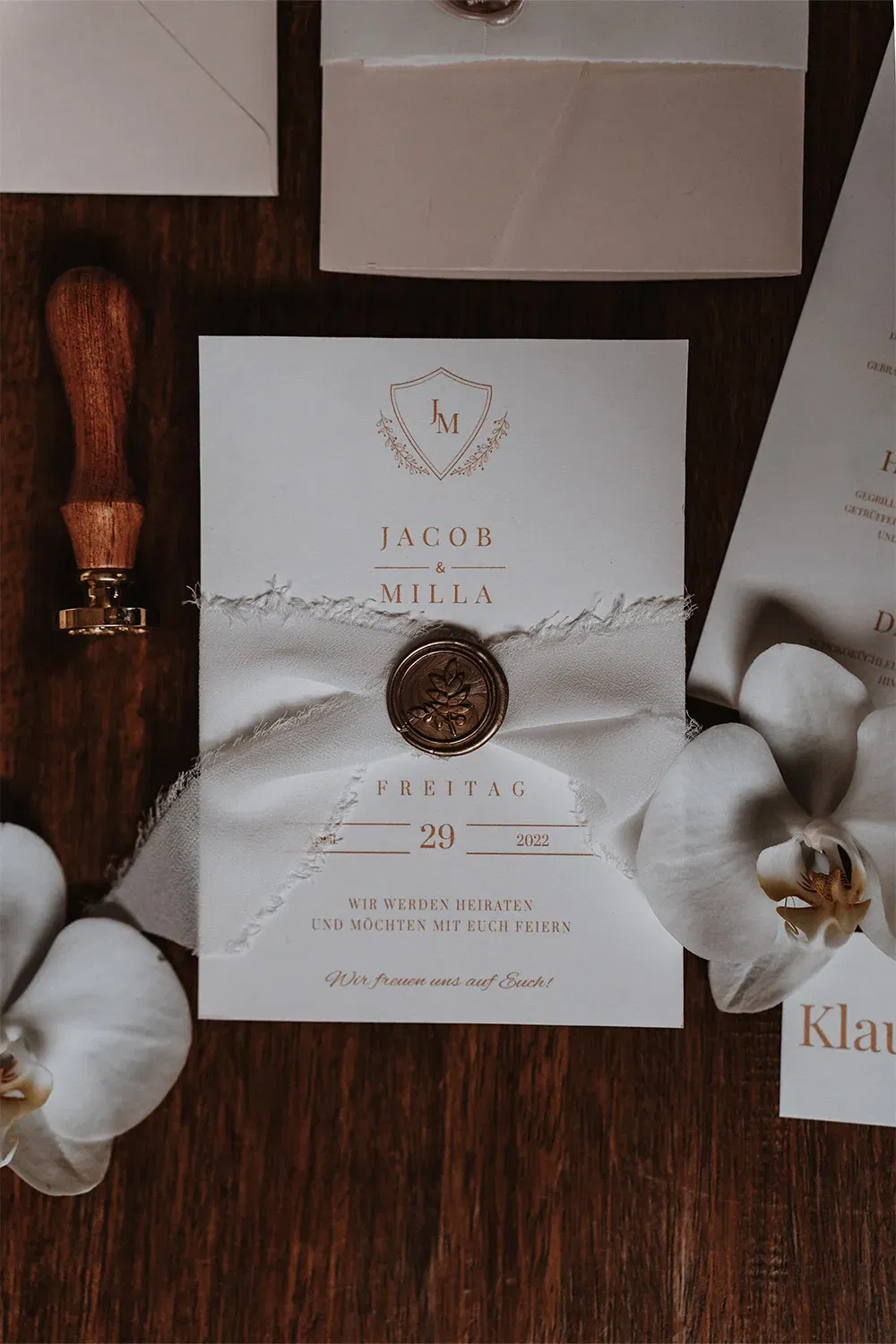 Einladung zur Hochzeit, umwickelt mit weißem Band und einem Wachssiegel, stilvoll auf einem Holztisch arrangiert, begleitet von Orchideen.