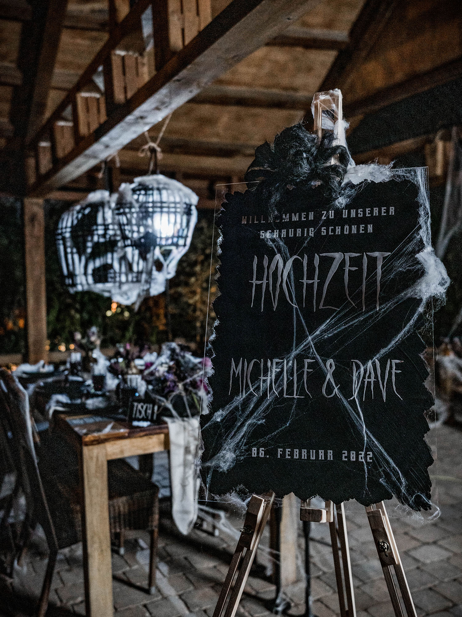 Schild mit den Namen "Michelle & Dave", umgeben von Spinnweben, kündigt eine düster-romantische Hochzeit an. Im Hintergrund ein gedeckter Tisch im Gothic-Design.