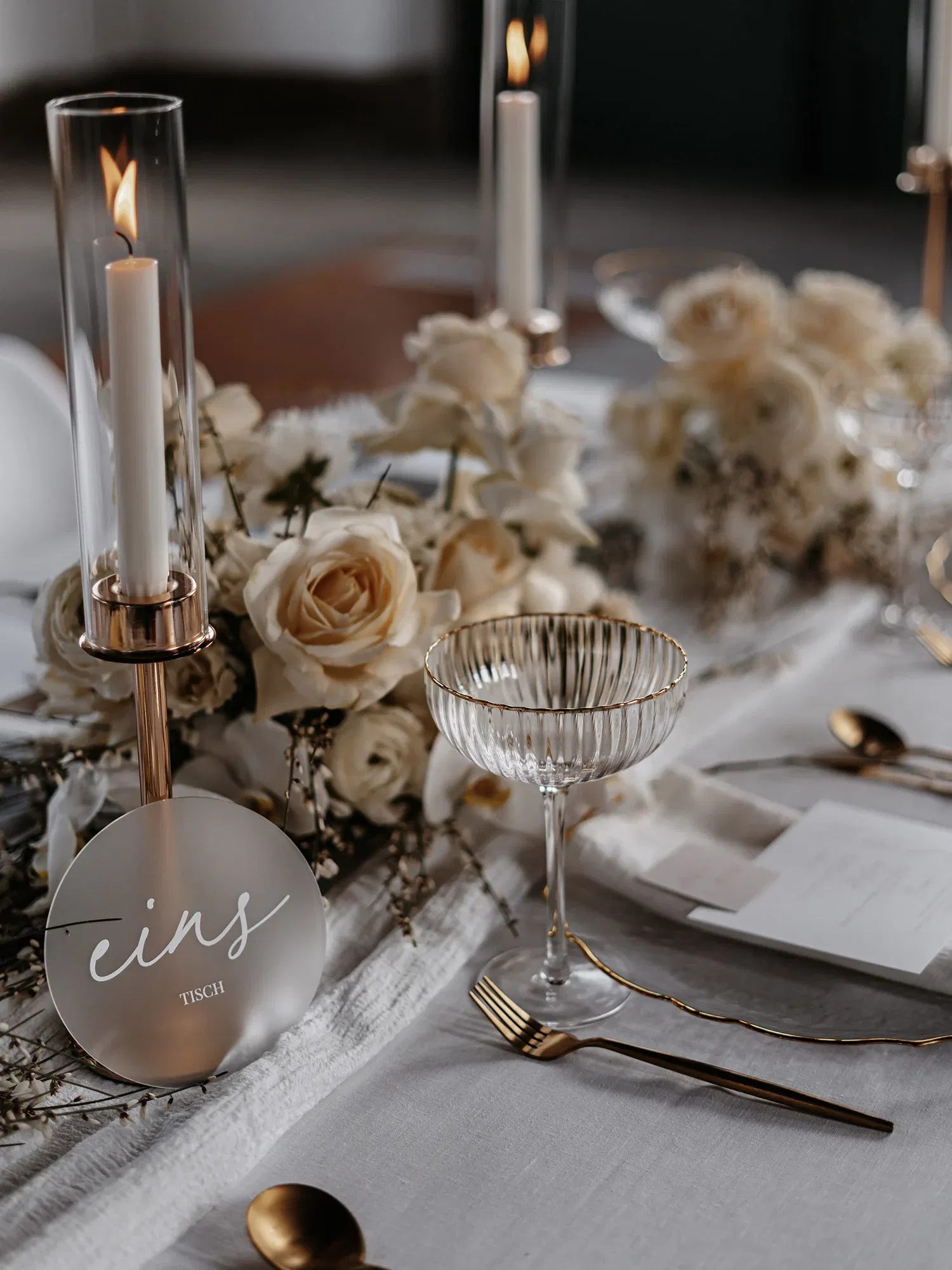 Eine feine Tischdekoration für eine Hochzeit, mit hellen Blumen, goldenen Akzenten und einer sanft flackernden Kerze in einem schmalen Glasgefäß, neben einer Tischnummer aus Acryl.