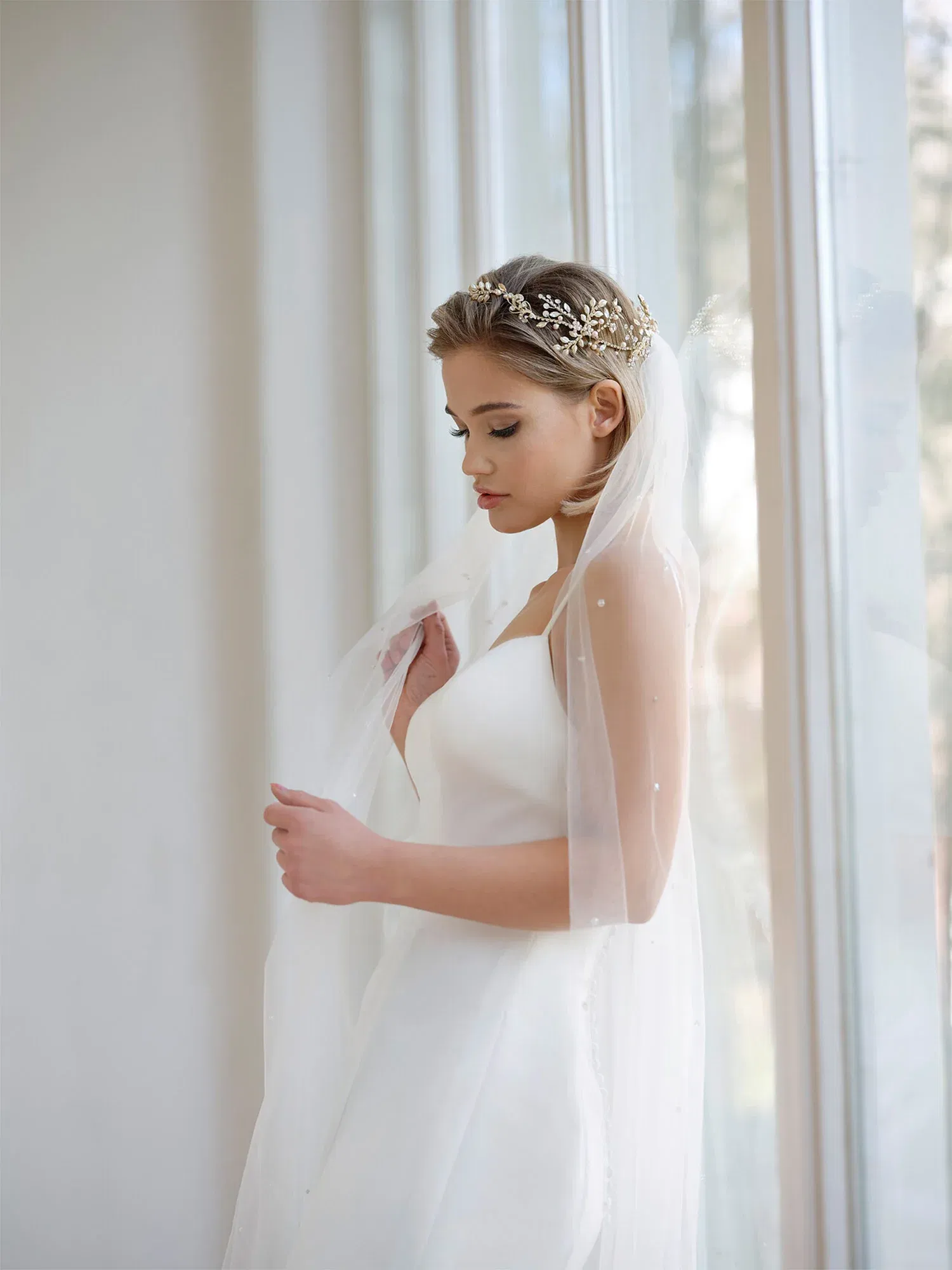 Seitliche Aufnahme einer Braut in einem schlichten Hochzeitskleid, die eine goldene Tiara und einen Schleier trägt, während sie an einem großen Fenster steht.