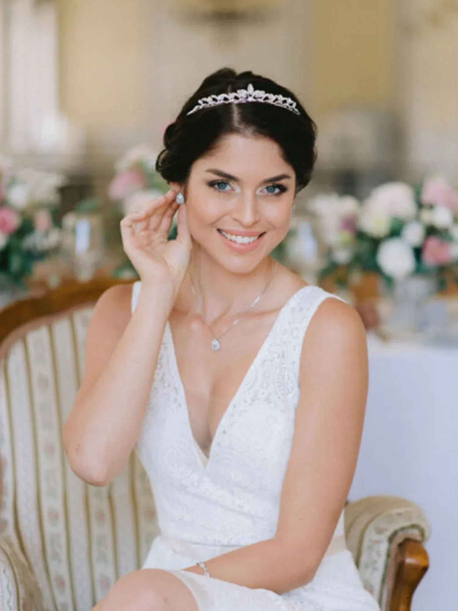 Lächelnde Braut in einem weißen Spitzenkleid sitzt auf einem klassischen Stuhl, gekrönt mit einer funkelnden Tiara und dezentem Schmuck.