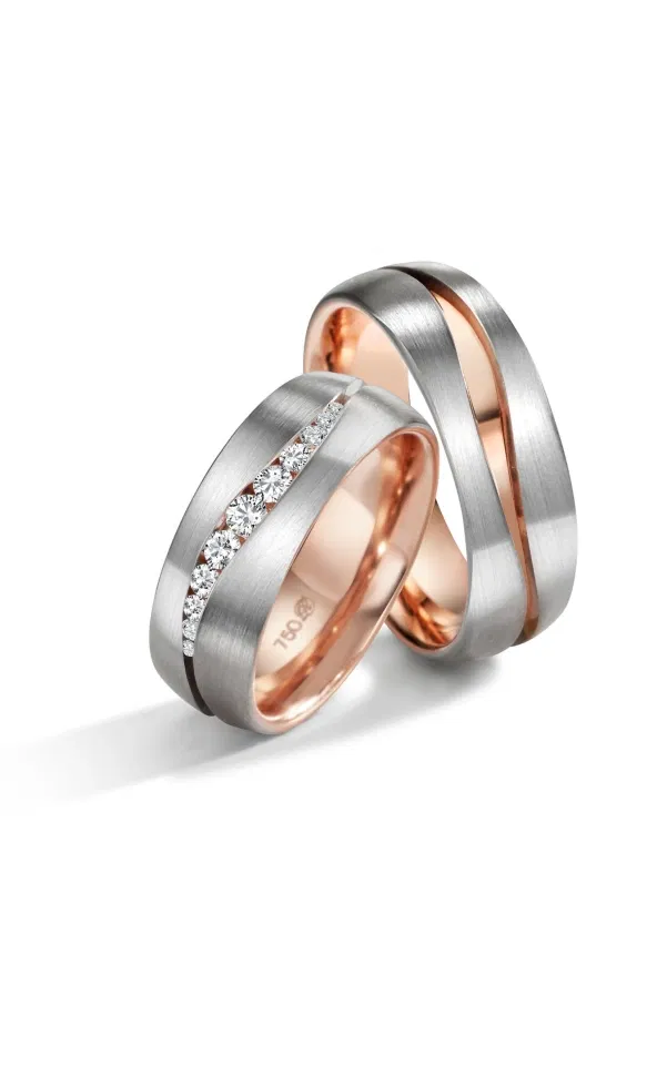 Zwei moderne Eheringe aus mattiertem Weißgold mit warmem Roségold-Innenband, einer davon mit einer schräg eingefassten Reihe funkelnder Diamanten, stilvoll vor hellem Hintergrund fotografiert.