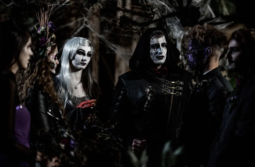 Hochzeitsgäste im Gothic-Stil, geschminkt mit Halloween-Motiven, stehen in einer düsteren Zeremonie zusammen. Die Szene ist von dunkler, mysteriöser Atmosphäre geprägt.